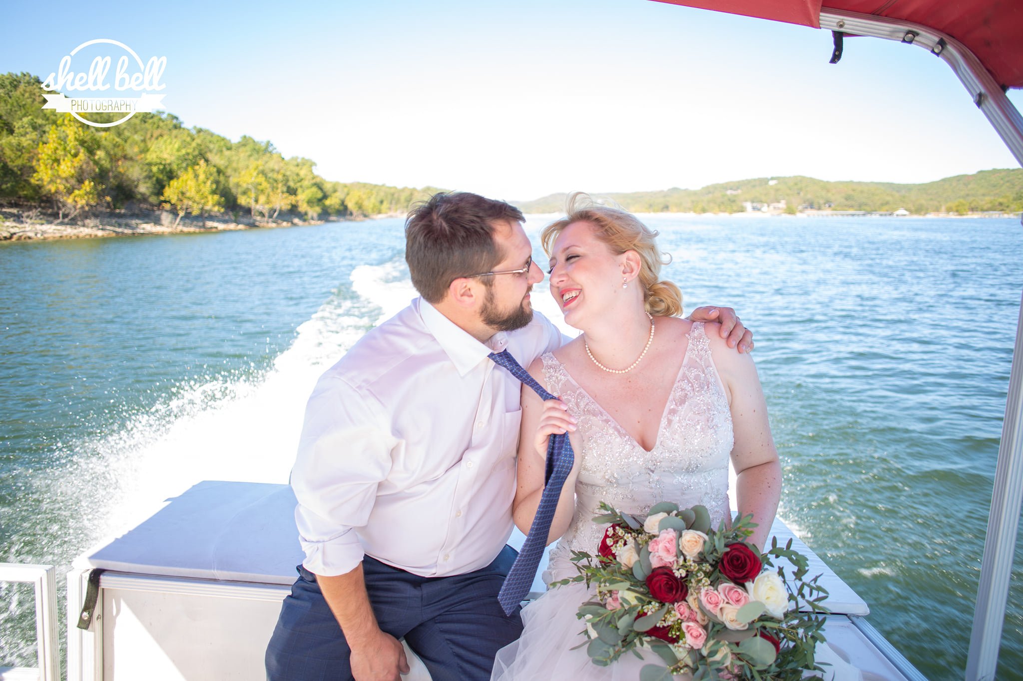 A newlywed couple sits close together on a boat, smiling and sharing a moment. They are on a lake with forested hills in the background, under a red canopy.