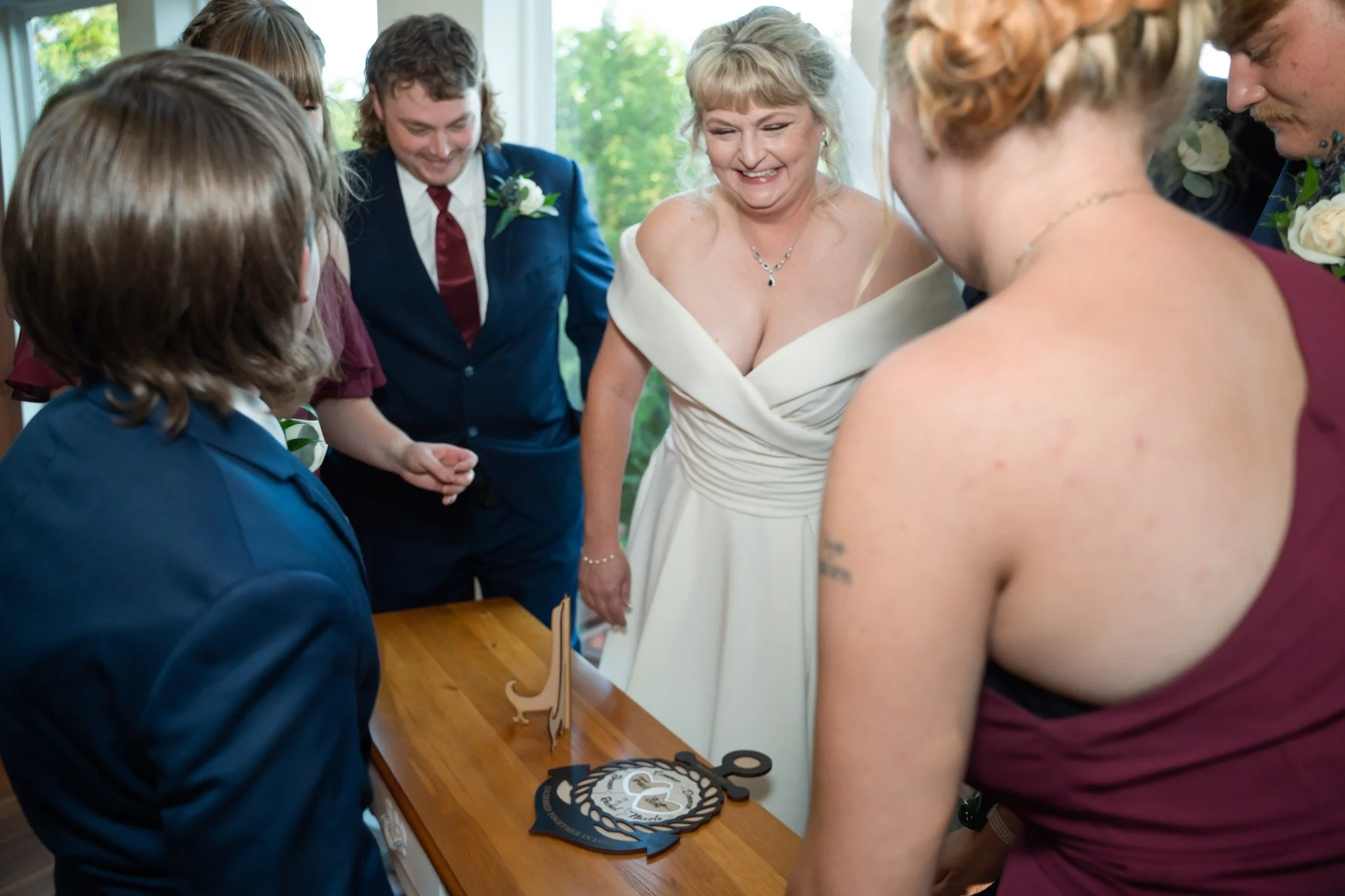 A bride and groom along with their wedding party looking down at a table with a wedding cake and decorative plaques, in a well-lit room with large windows and greenery outside.