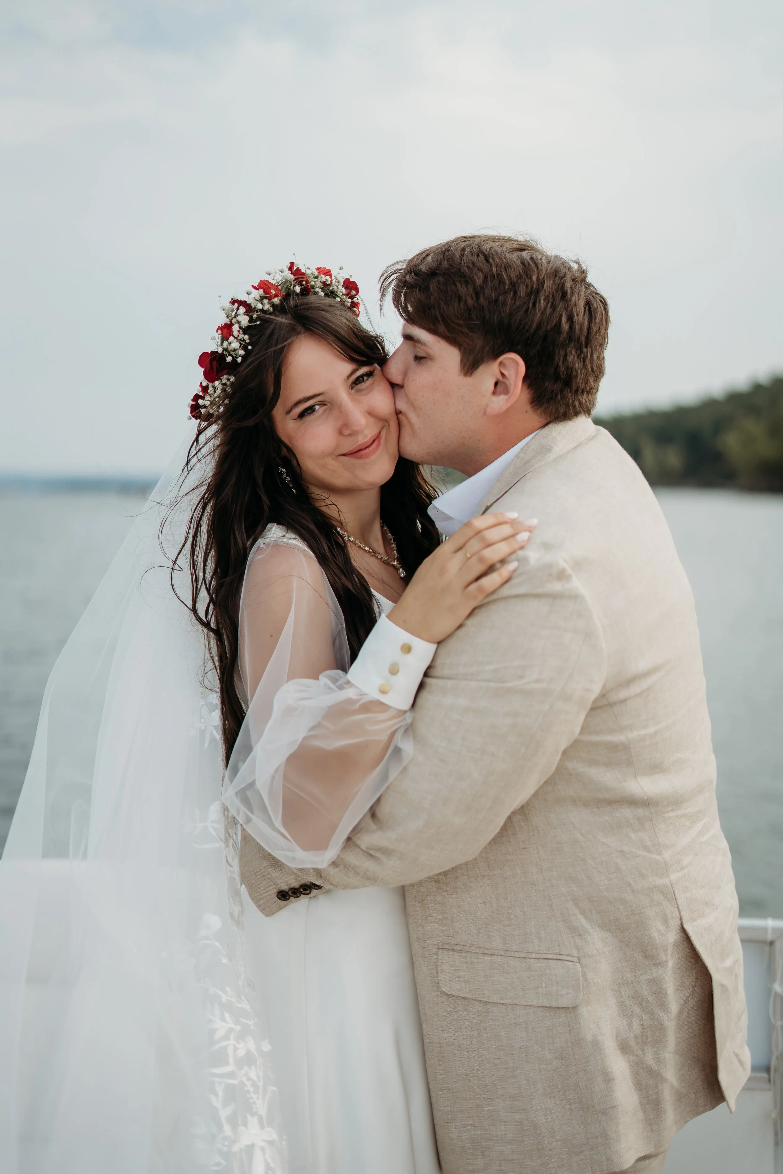 A bride and groom embrace outdoors near a body of water, with the groom kissing the bride on the cheek. The bride wears a white wedding dress, a floral crown, and has long dark hair. The groom wears a beige suit.
