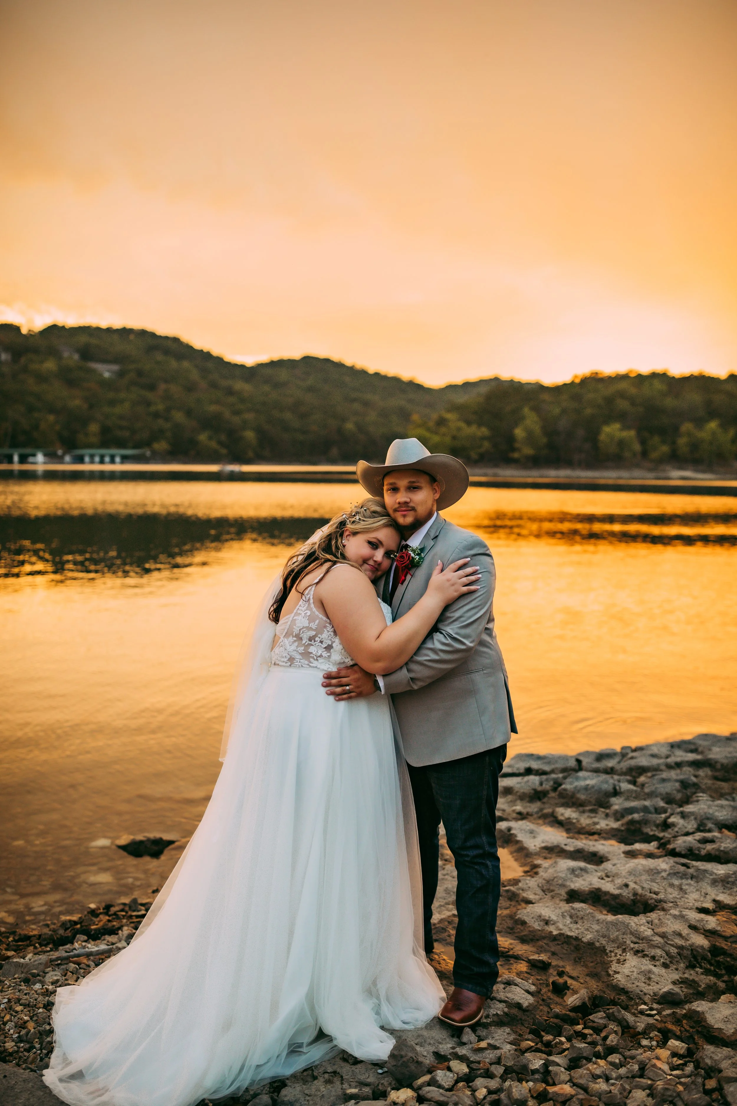 A bride and groom embracing by a lake during sunset, with mountains and a colorful sky in the background.