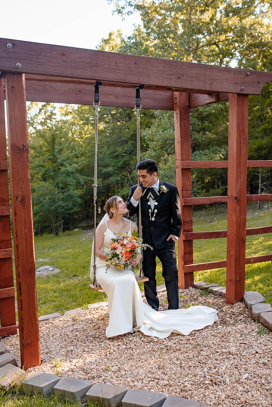 A bride and groom in wedding attire on a wooden swing set outdoors, with trees and greenery in the background.