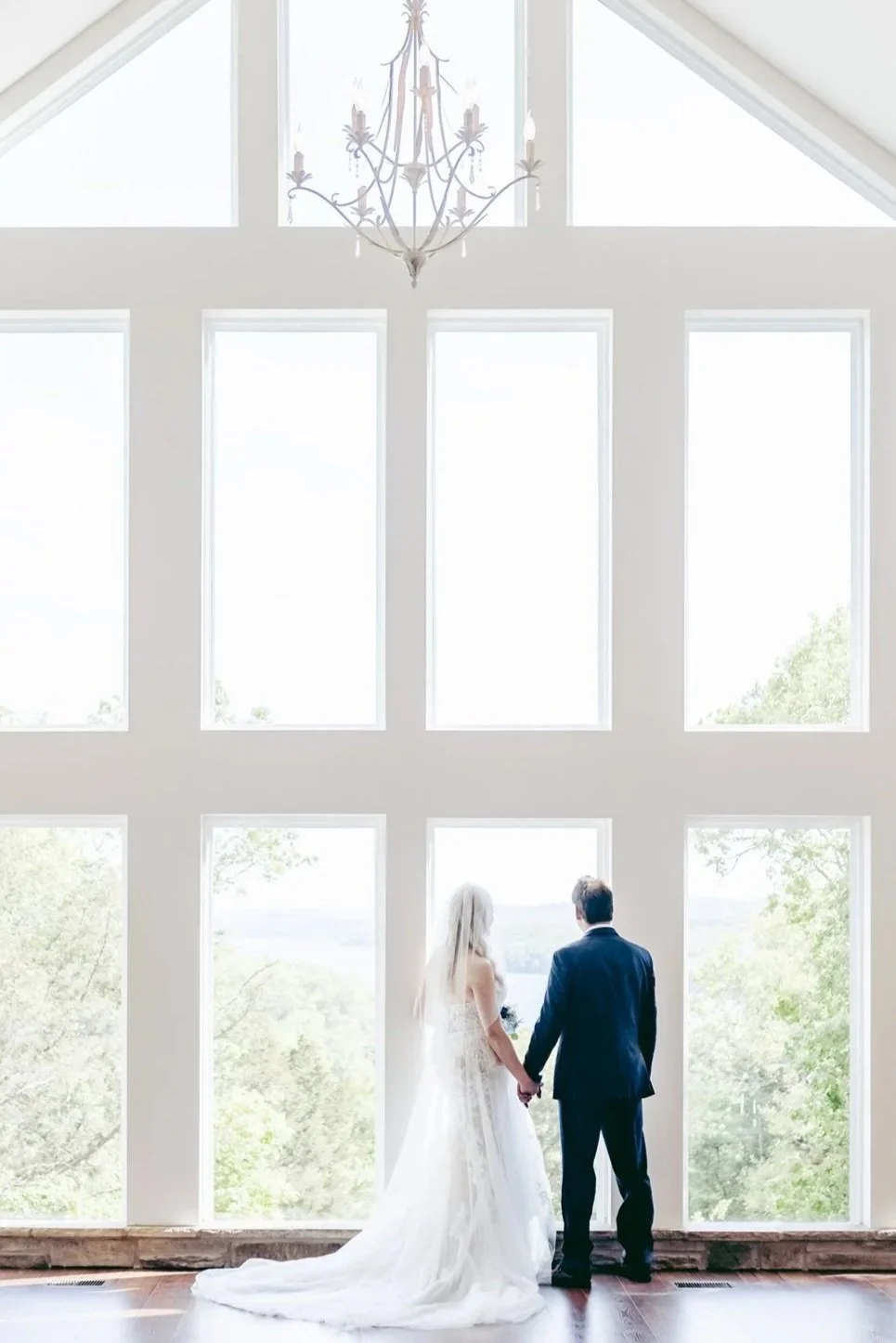 A bride and groom holding hands, standing in front of large windows inside a bright, modern room with a chandelier hanging from the ceiling. The bride is wearing a white wedding dress with a long train, and the groom is dressed in a dark suit.