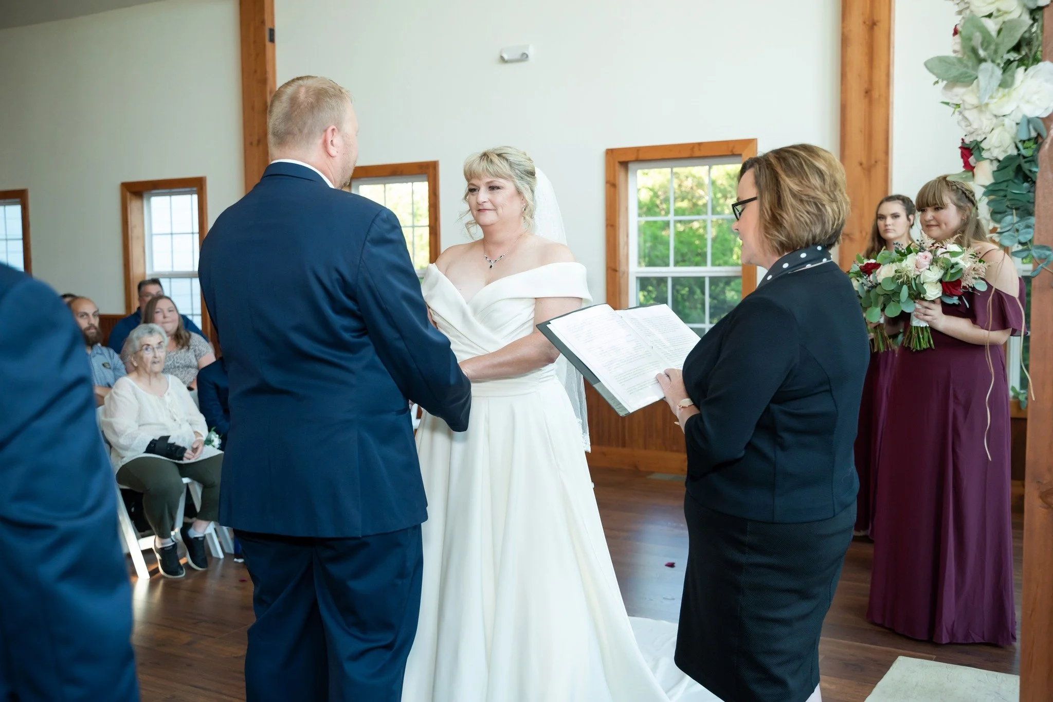 A couple getting married during a ceremony in a wooden-floored room with large windows, surrounded by an audience and a woman officiant reading from a booklet, with bridesmaids holding flower bouquets in the background.