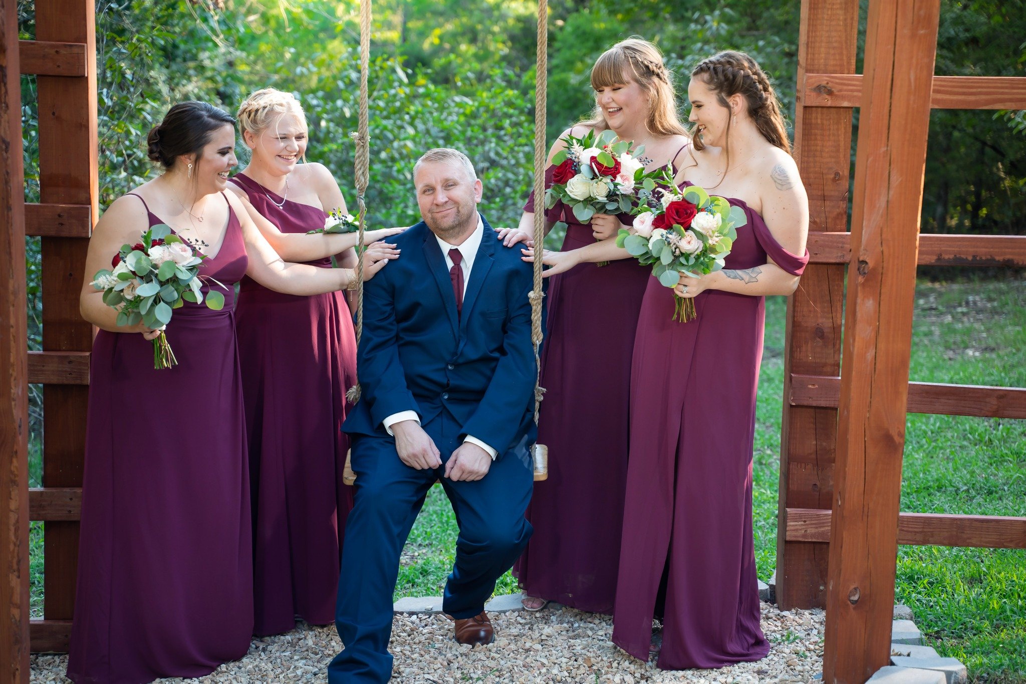 A man sitting on a swing surrounded by four women in purple dresses holding bouquets of flowers, outdoors in a wooded area.