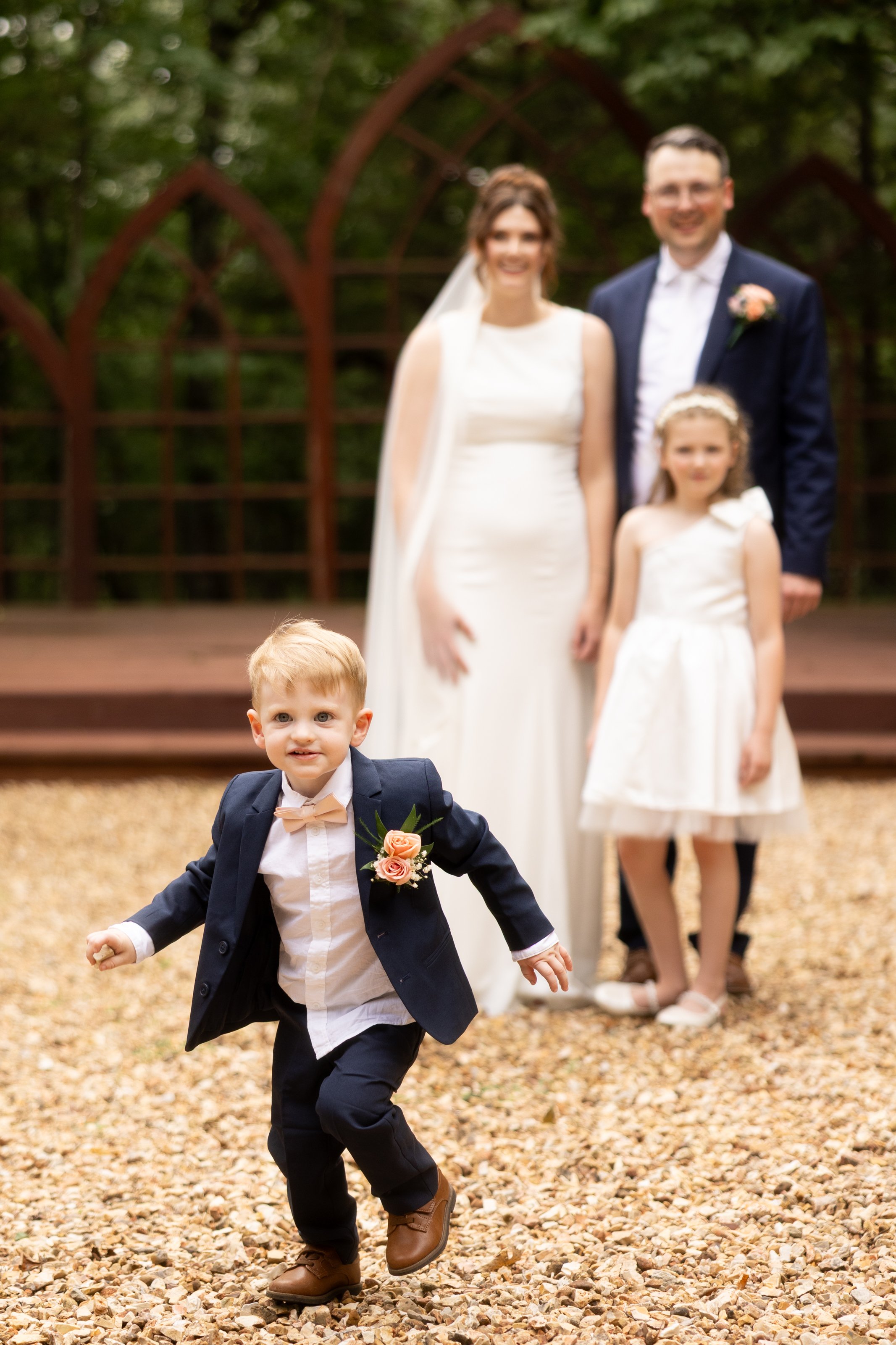 A young boy in a navy suit and pink bow tie is running towards the camera, with a wedding party in the background. The wedding party includes a bride and groom, and two young girls, all standing outdoors on a gravel path surrounded by trees and a decorative wooden arch.