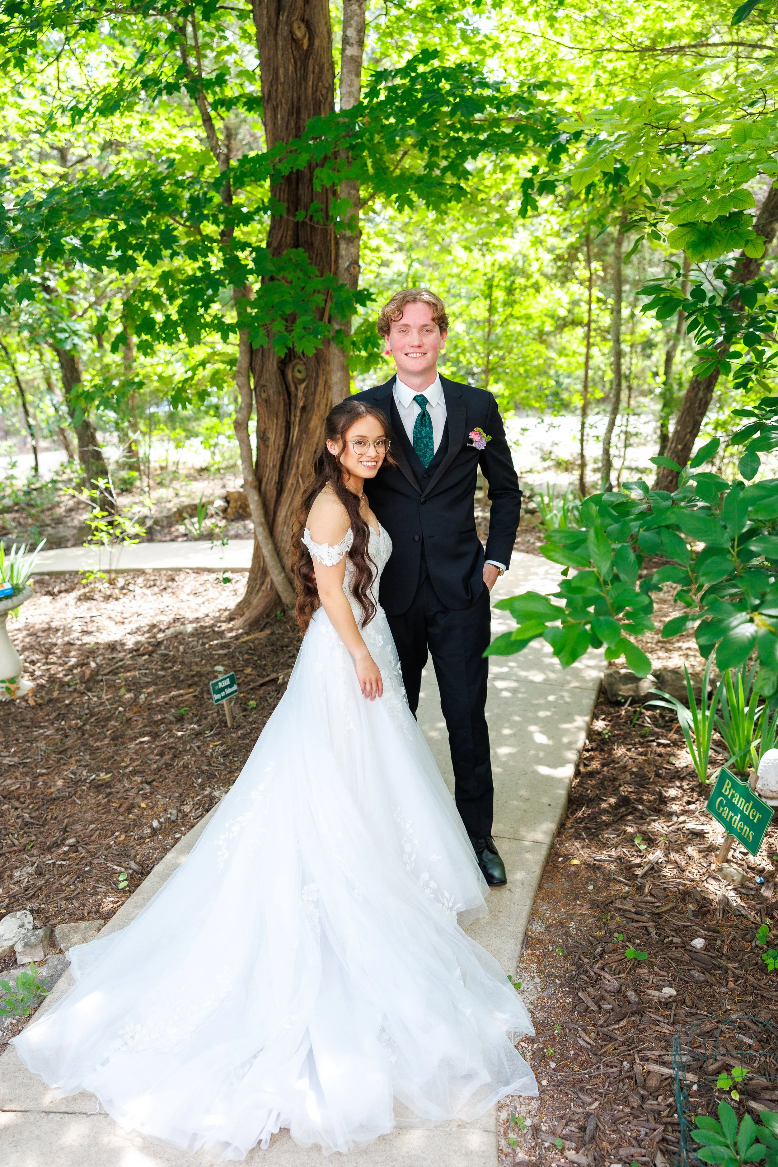 A couple dressed in wedding attire standing on a garden path surrounded by green trees and foliage. The bride in a white wedding gown with a long train and the groom in a black suit with a vest and tie.