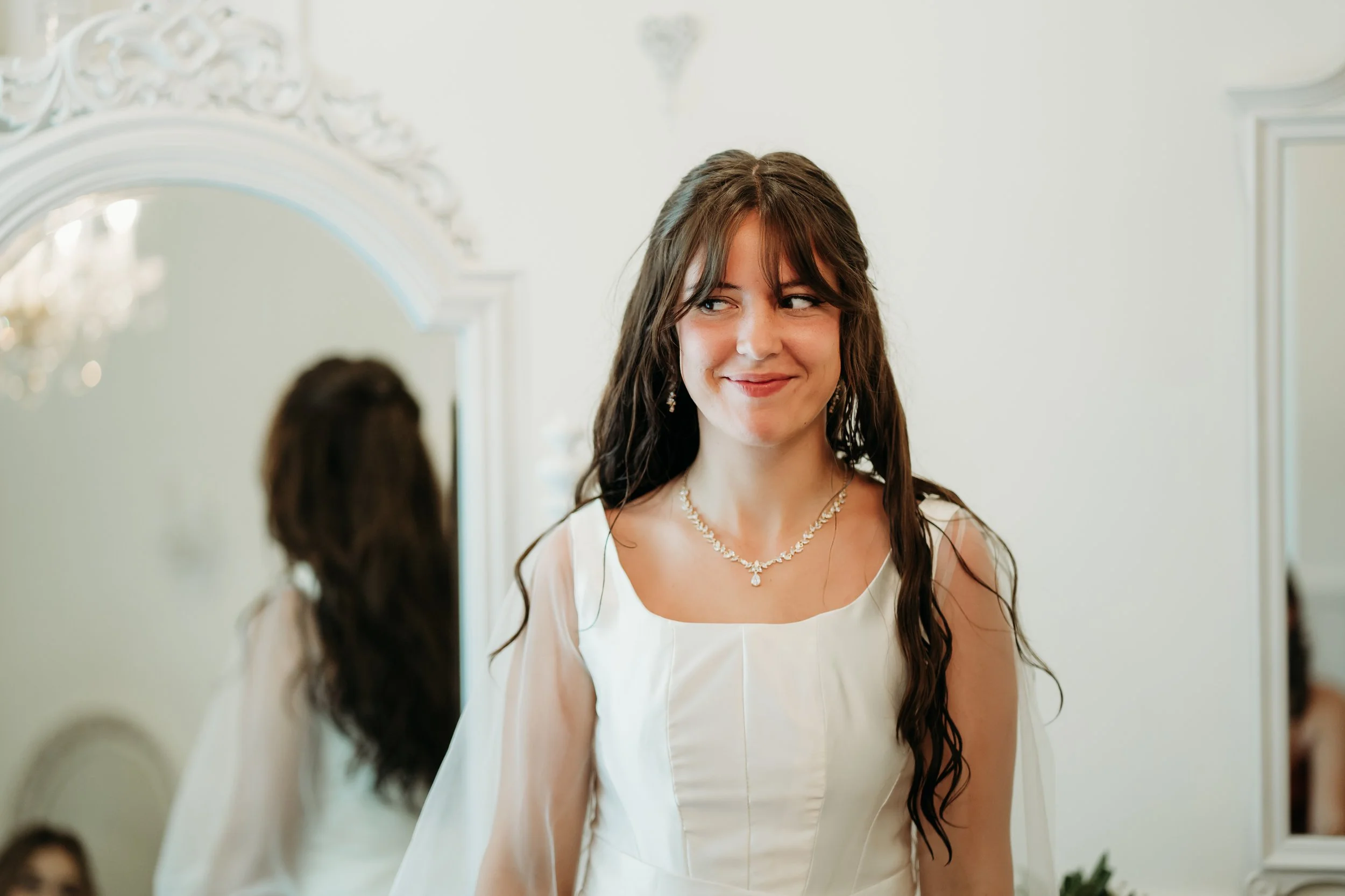 Young woman with long, dark hair wearing a white dress and necklace, looking at herself in a mirror during a wedding or special occasion.