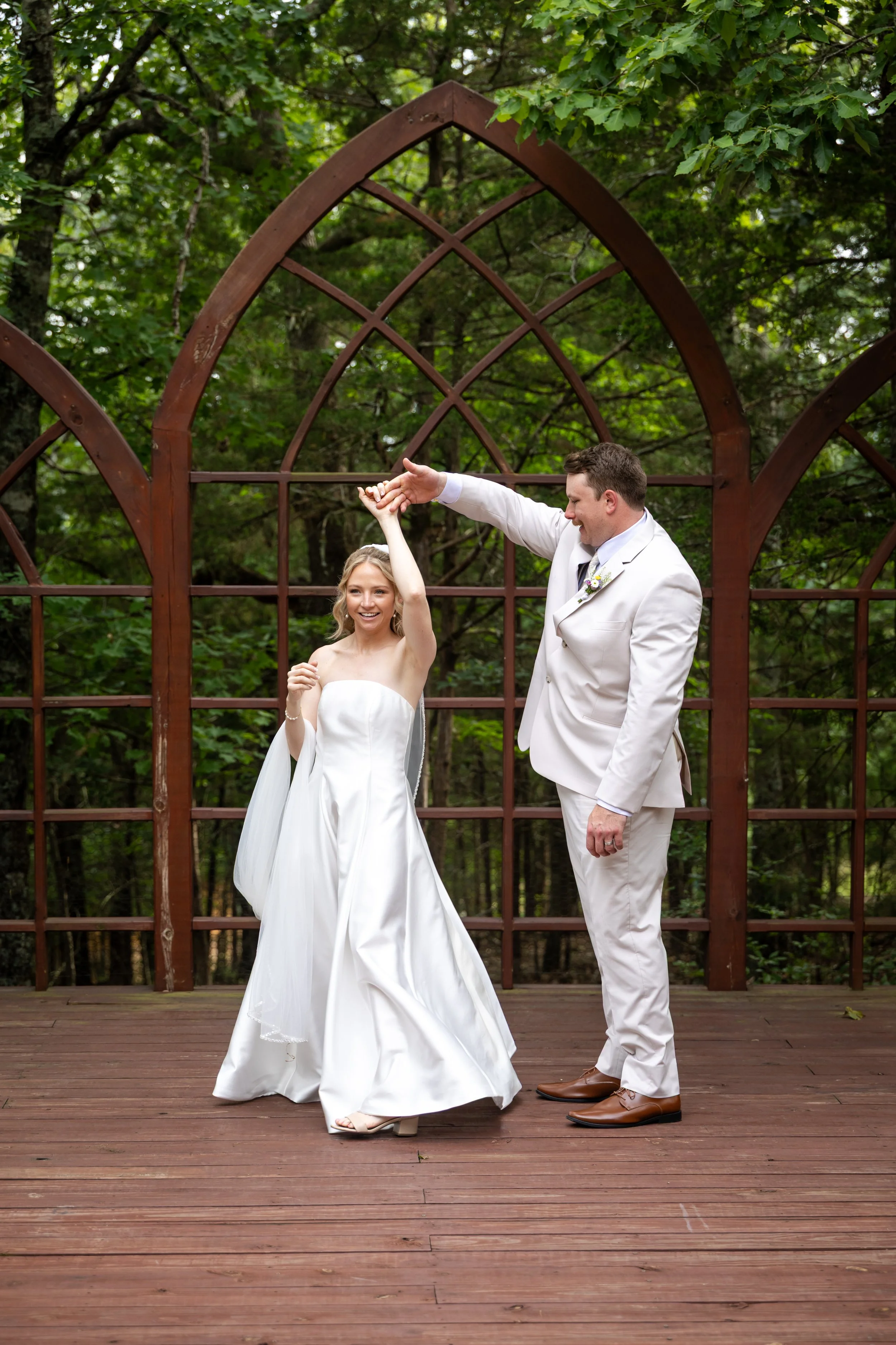 A newlywed couple, the bride in a strapless white wedding gown and the groom in a light-colored suit, dance outdoors under a wooden arbor with green trees in the background.