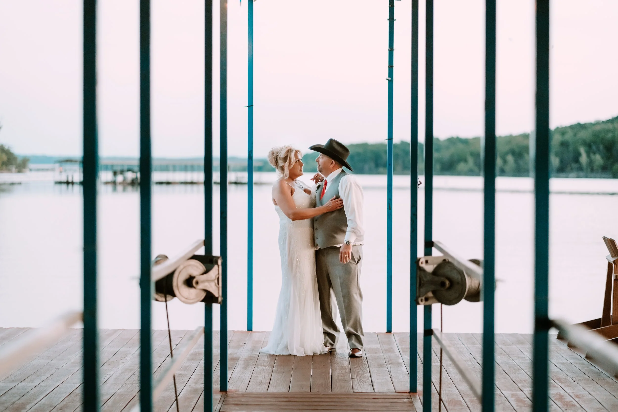 A bride and groom stand close together on a wooden dock near a body of water, seen through blue metal bars. The bride wears a white wedding dress, and the groom wears a gray vest, white shirt, red tie, and a black cowboy hat. The background features 