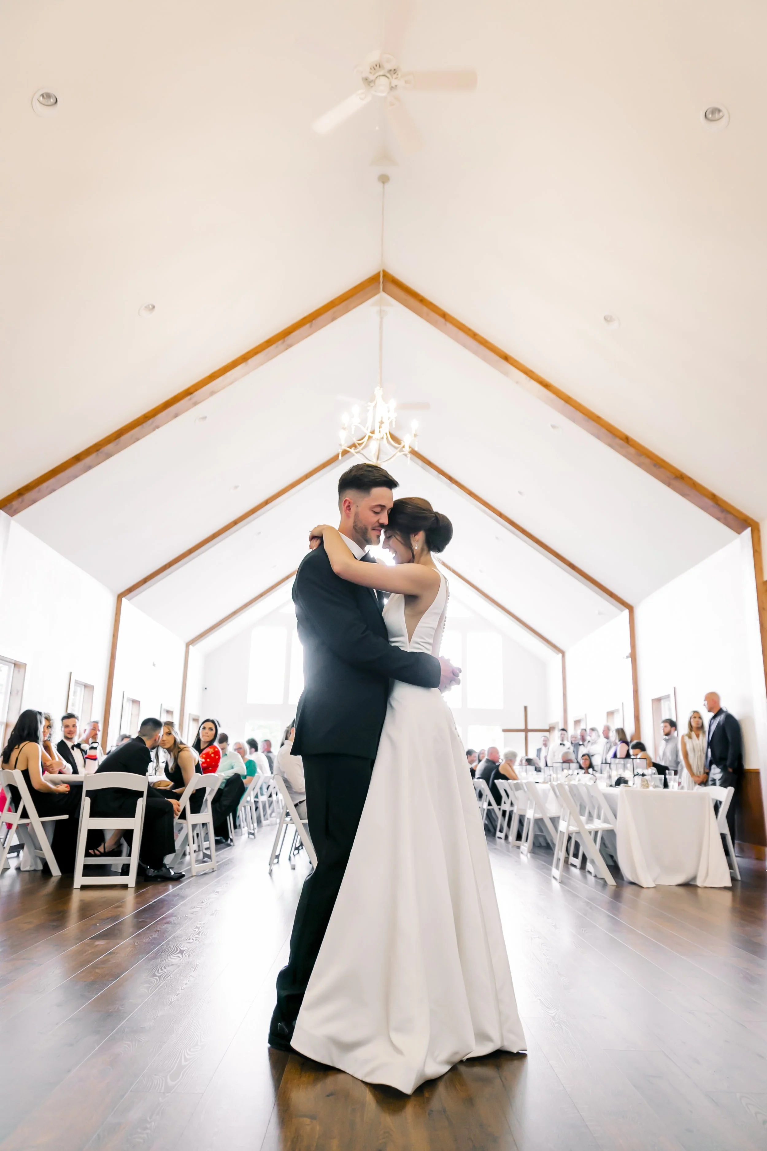 A bride and groom embrace and dance in a bright, spacious wedding hall with wooden accents, chandeliers, and guests seated at tables.