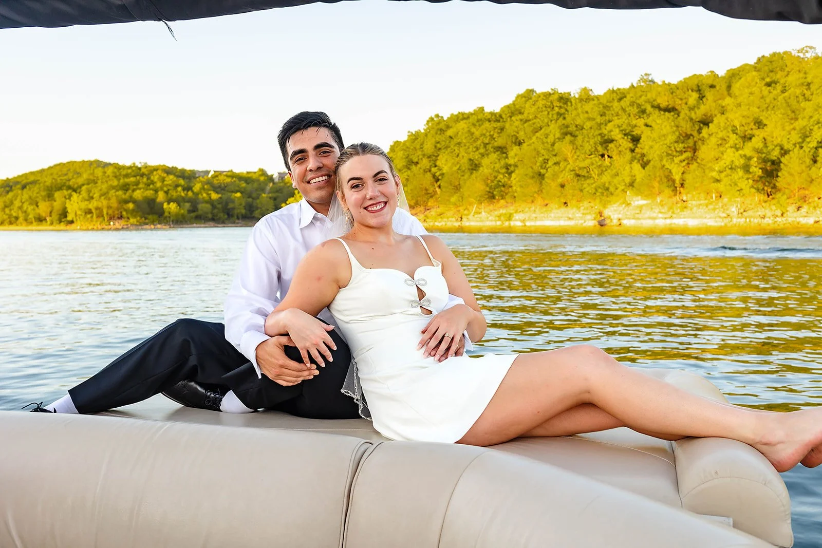 Smiling couple in wedding attire sitting on a boat near the water, with green trees and hills in the background during sunset.