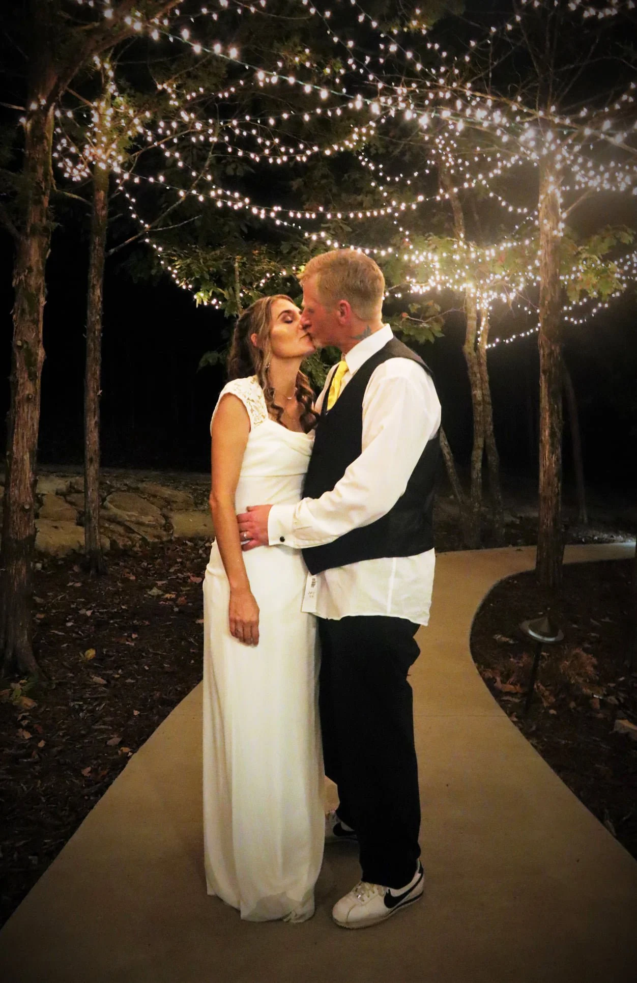 A bride and groom share a kiss under string lights hanging from trees at night, standing on a curved pathway outdoors.