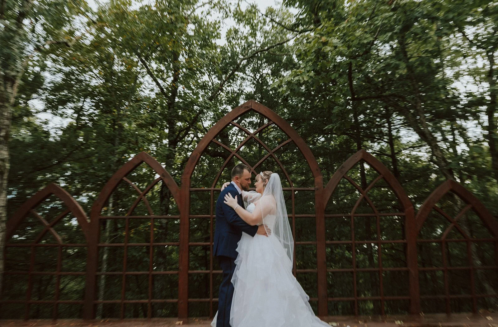 A newlywed couple embraces in front of a rusted, gothic-style metal trellis in a forest setting.