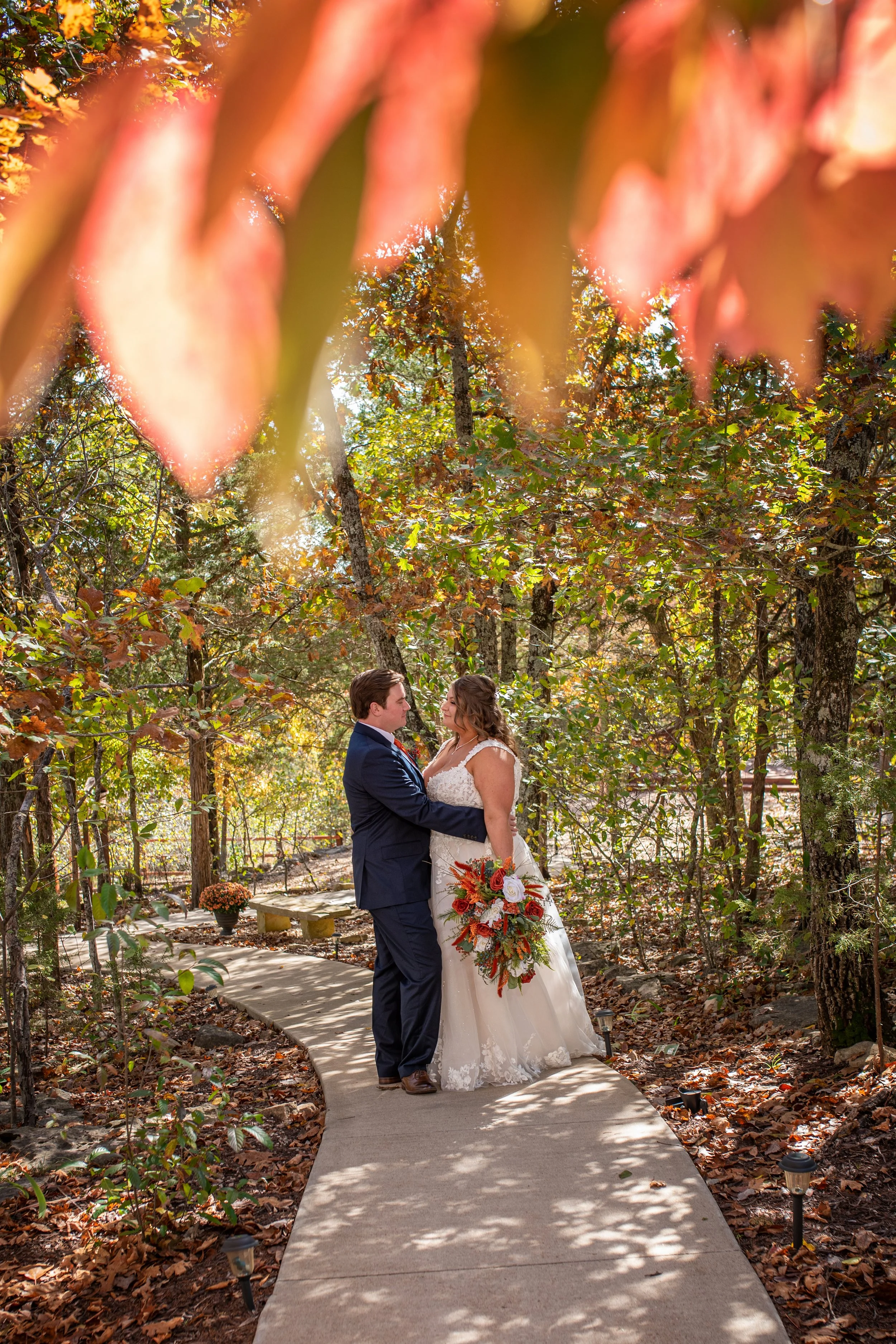 A bride and groom stand on a garden pathway surrounded by trees with fall leaves, gazing into each other's eyes. The bride holds a bouquet of orange, red, and white flowers.