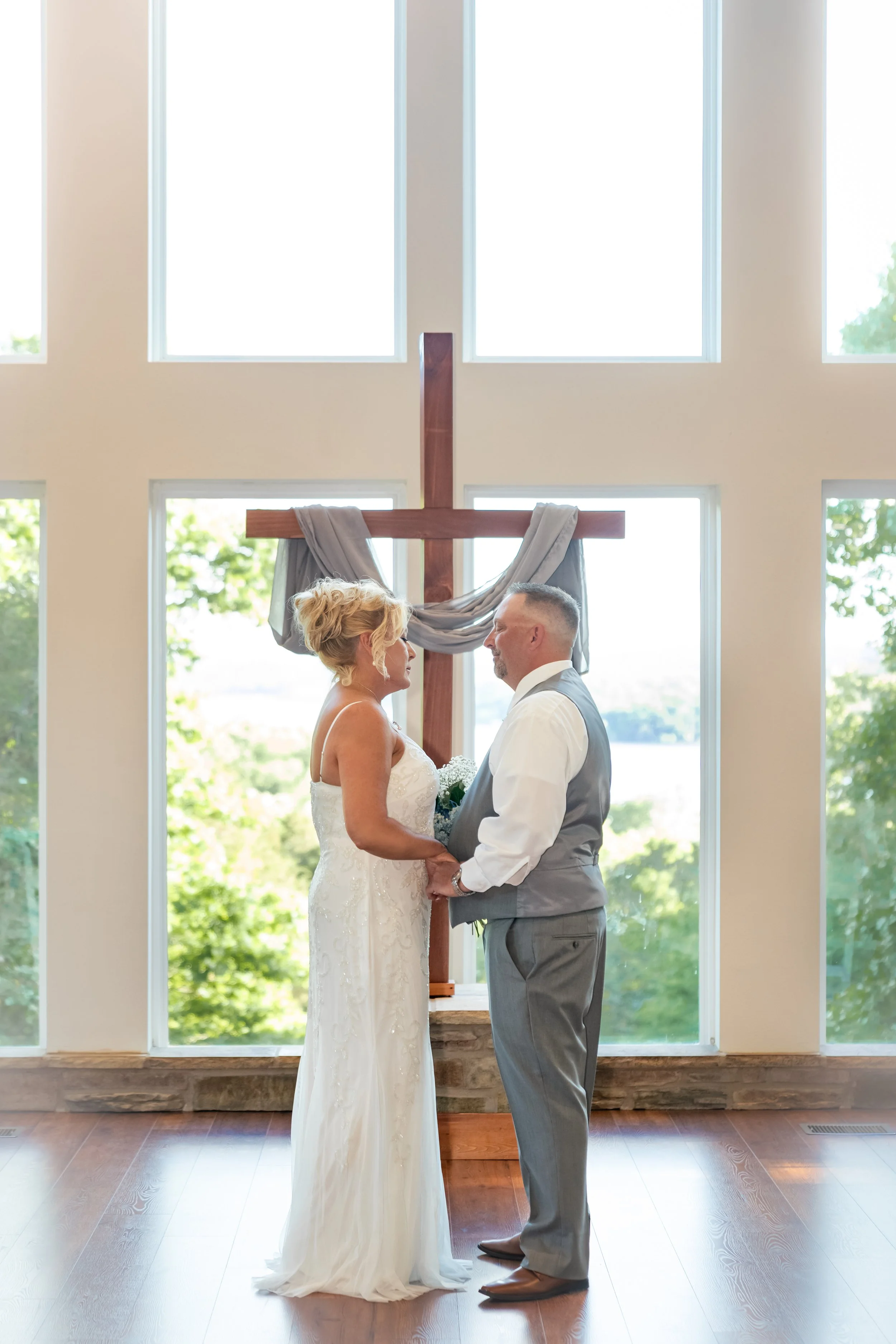 A bride and groom standing facing each other inside a church, holding hands, with a large cross behind them and tall windows letting in natural light.