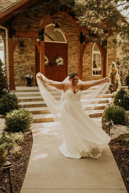 Bride in a white wedding gown dancing on a pathway outside a rustic brick house with stairs and greenery.