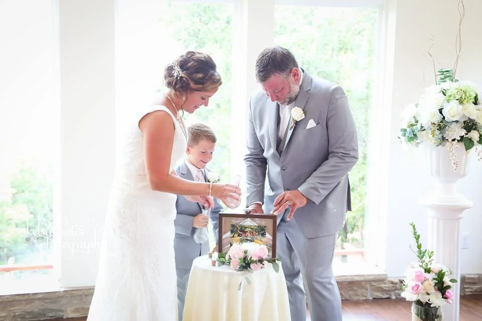 A wedding ceremony with a bride, groom, and a young boy, all smiling and looking at a framed picture on a table decorated with pink and white flowers.