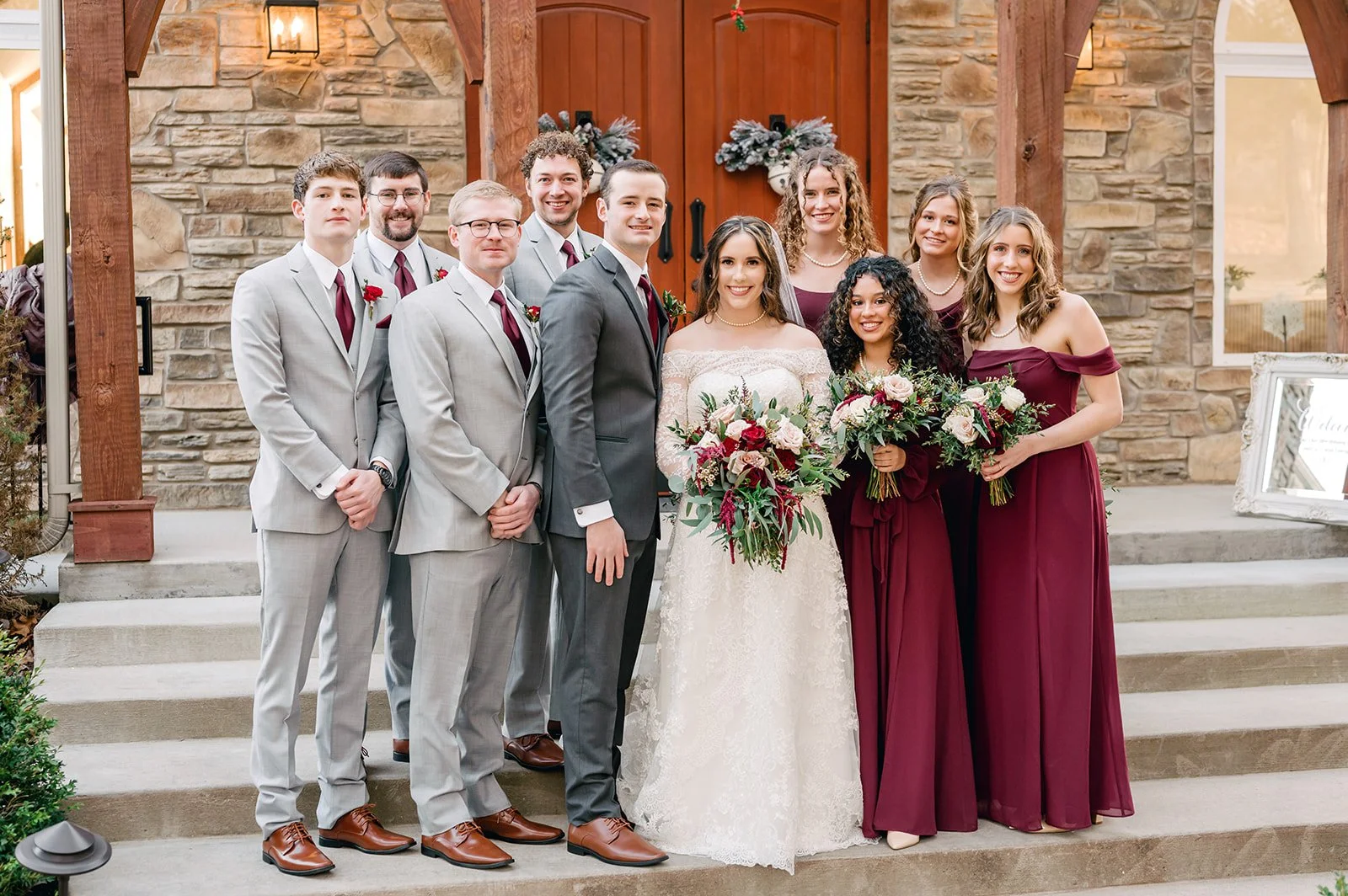 A wedding party standing on steps outside a building with stone walls and wooden doors, dressed in formal attire. The bride is in a white lace gown holding a large bouquet, and the groom is in a gray suit with a white shirt and black tie. Seven bridesmaids in burgundy dresses and four groomsmen in light gray suits with burgundy ties are present.