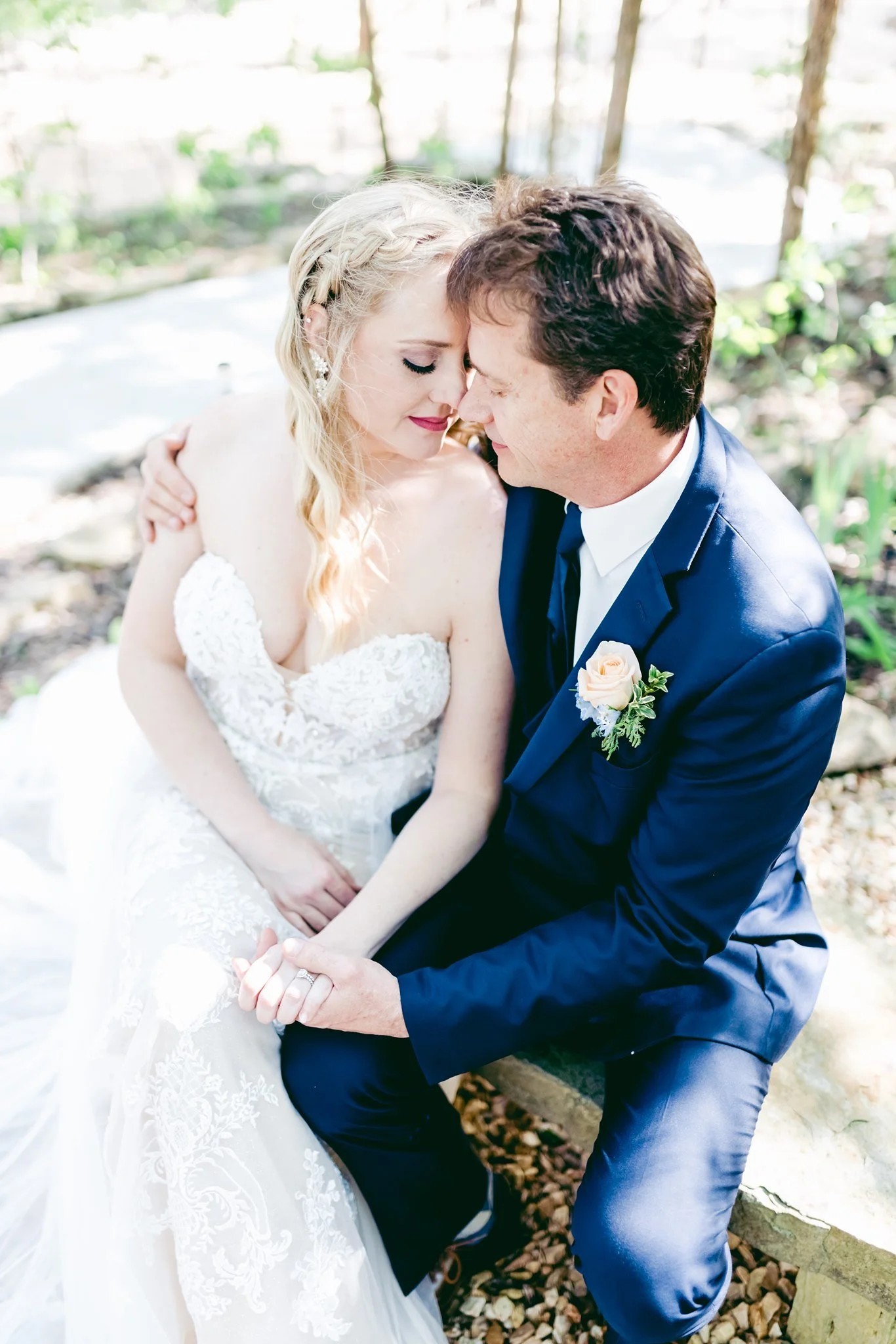 A bride and groom sitting close together outdoors, with their foreheads touching and eyes closed, sharing an intimate moment on their wedding day.