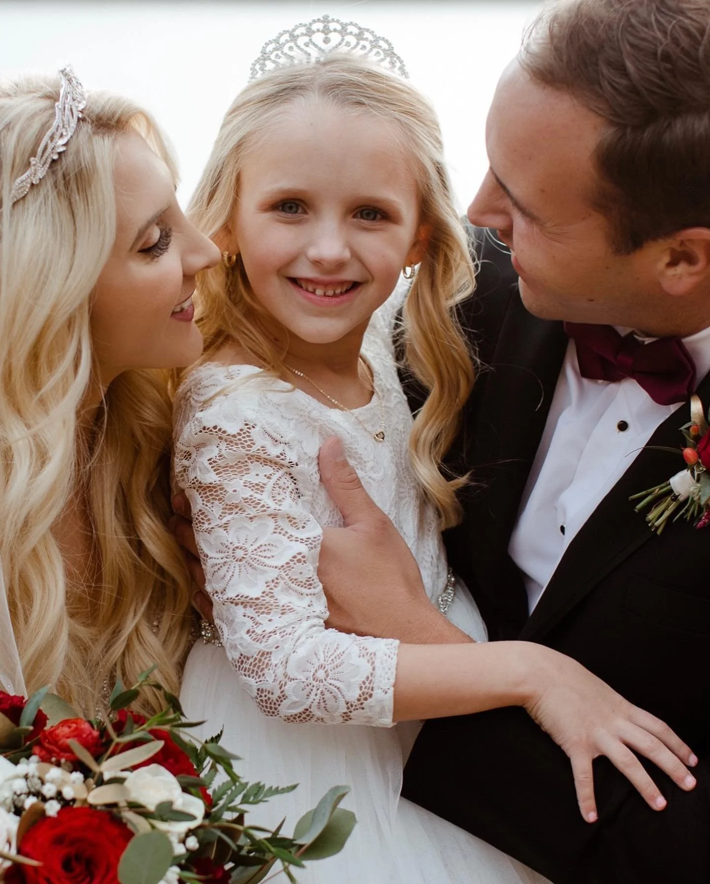 A young girl in a white lace dress with a tiara and earrings smiling at an adult woman with blond hair and a tiara and a man in a tuxedo with a red bowtie, all closely together at a wedding celebration.