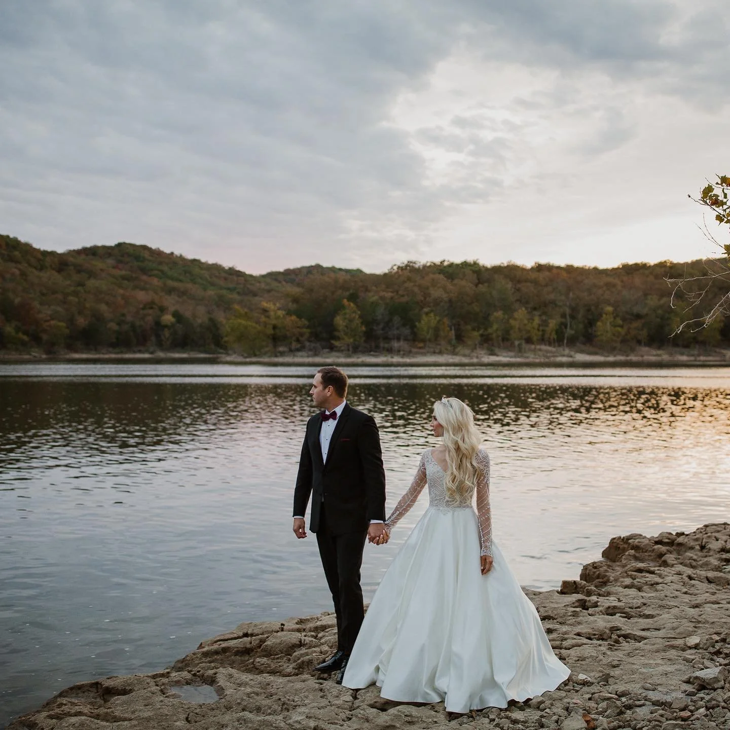 A bride and groom in wedding attire holding hands on a rocky riverbank at sunset, with a wooded hillside and cloudy sky in the background.