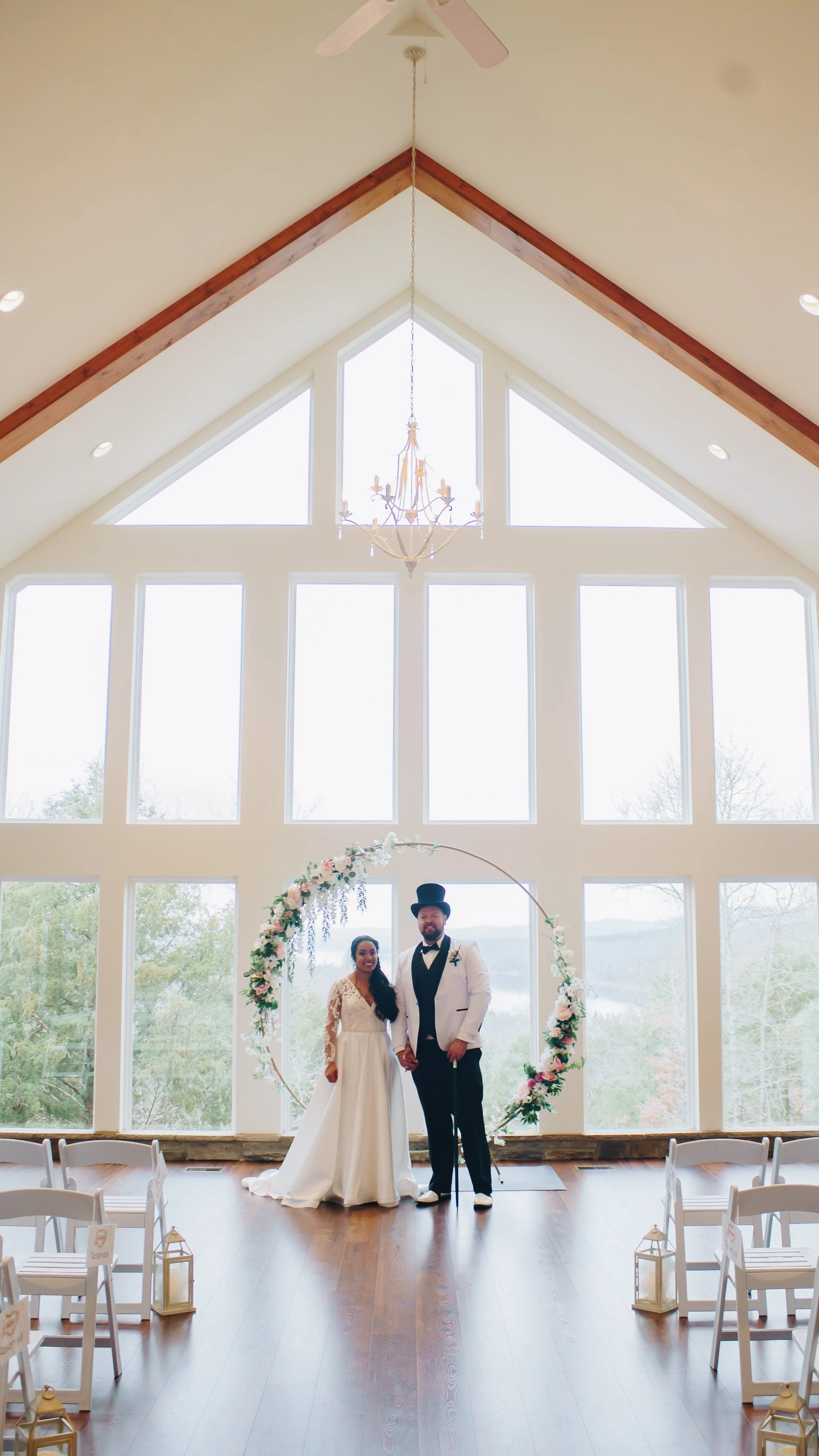 A bride and groom standing inside a light-filled wedding venue with large windows, a high vaulted ceiling with a chandelier, and an arch decorated with flowers behind them.