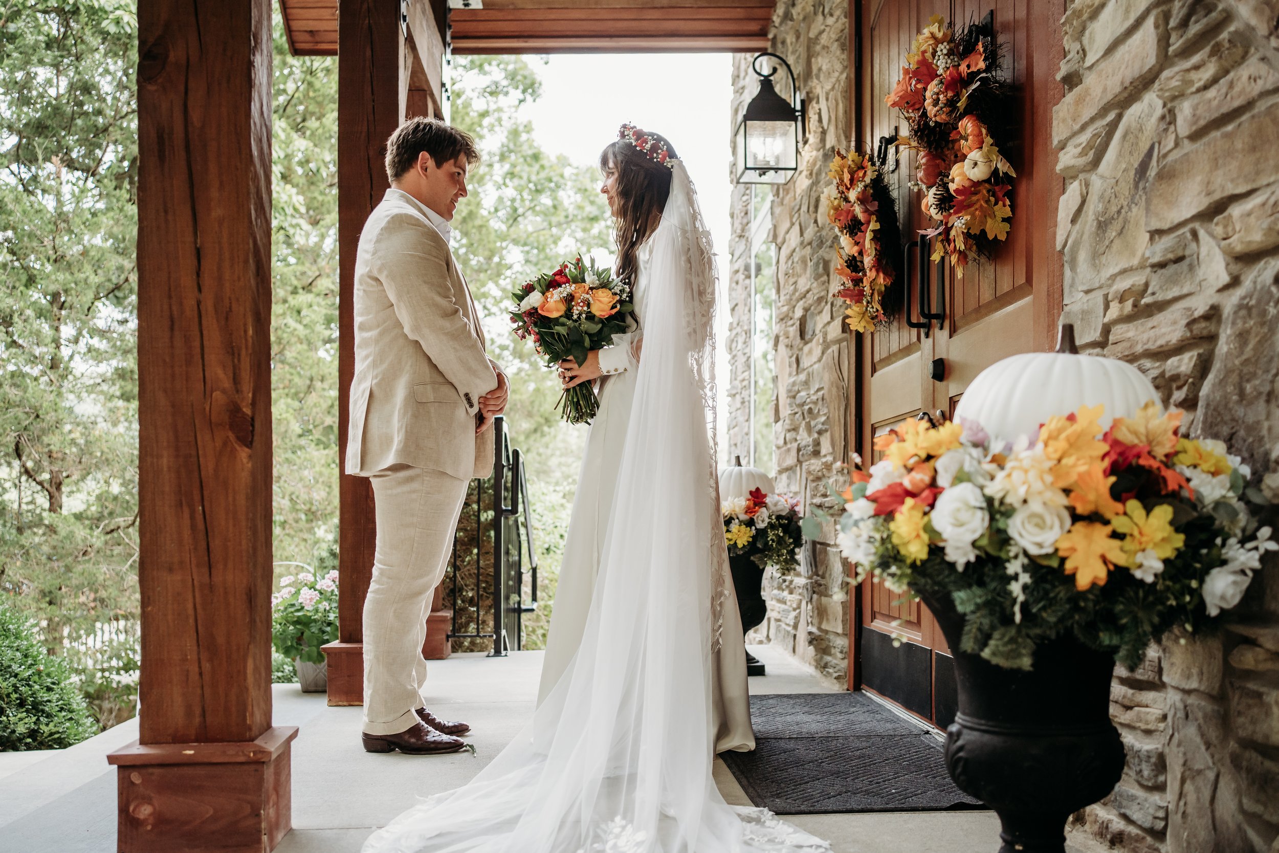 A bride and groom stand facing each other on a porch decorated for a wedding, with trees in the background. The bride is holding a bouquet and wearing a white wedding dress with a floral crown. The groom is dressed in a light-colored suit. Decor incl