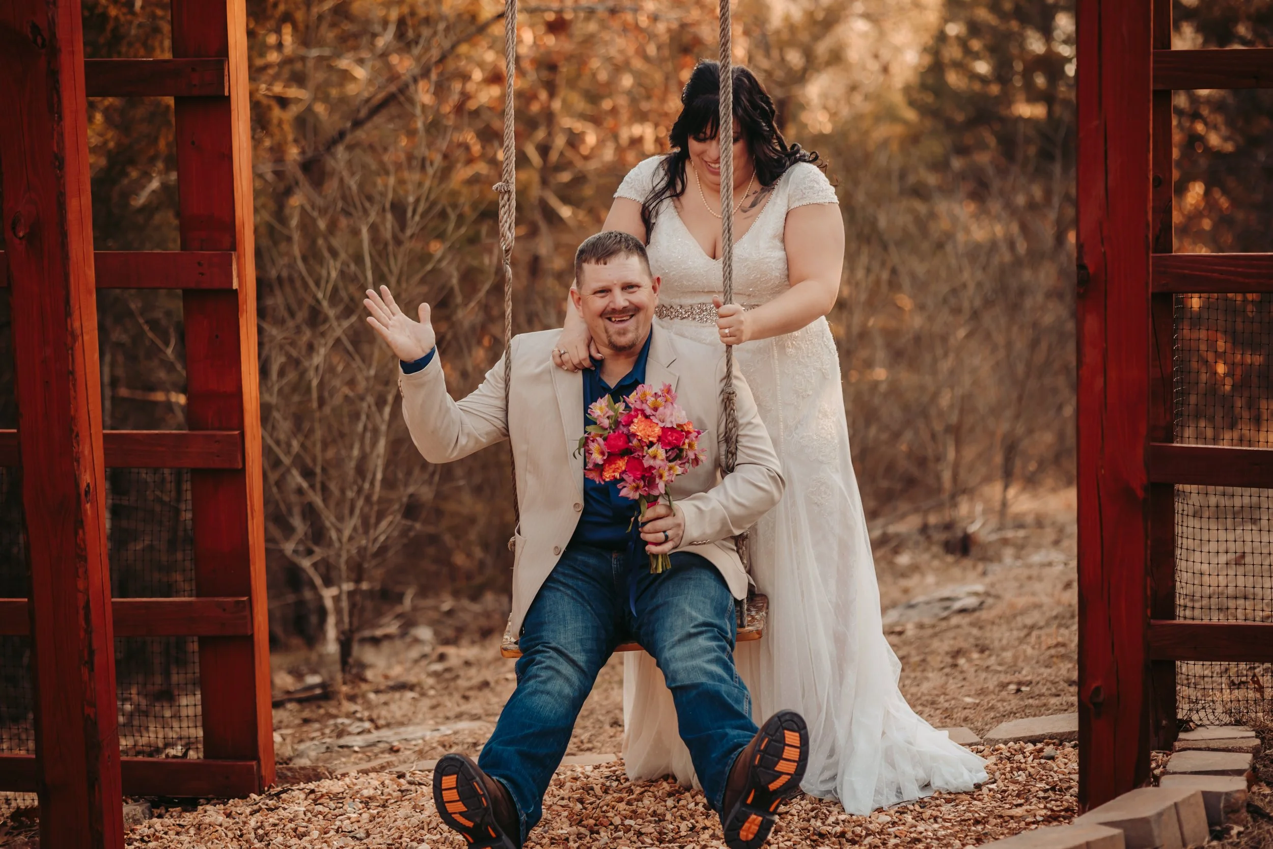 A woman in a wedding dress and a man in a beige blazer sitting on a swing, holding a bouquet, outdoors surrounded by fall trees.