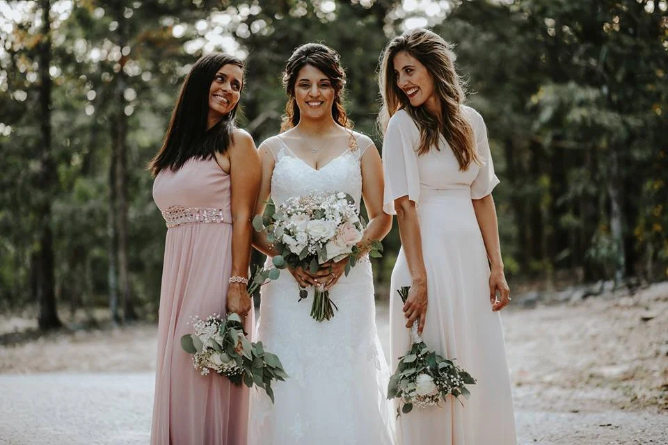Three women, one bride in a white wedding dress holding a bouquet, and two bridesmaids in pastel dresses holding smaller bouquets, standing outdoors among trees.