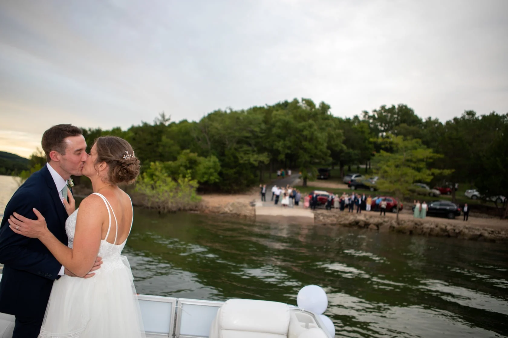A newlywed couple sharing a kiss on a boat during their wedding celebration, with guests gathered on a lakeshore in the background.