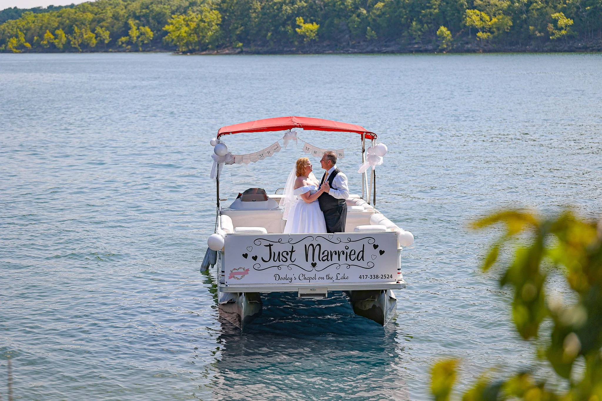 A boat on a lake with a couple celebrating their wedding, holding each other and smiling. The boat has a sign that reads "Just Married" and decorations, and a banner with "MR" and "MRS" hanging above them.