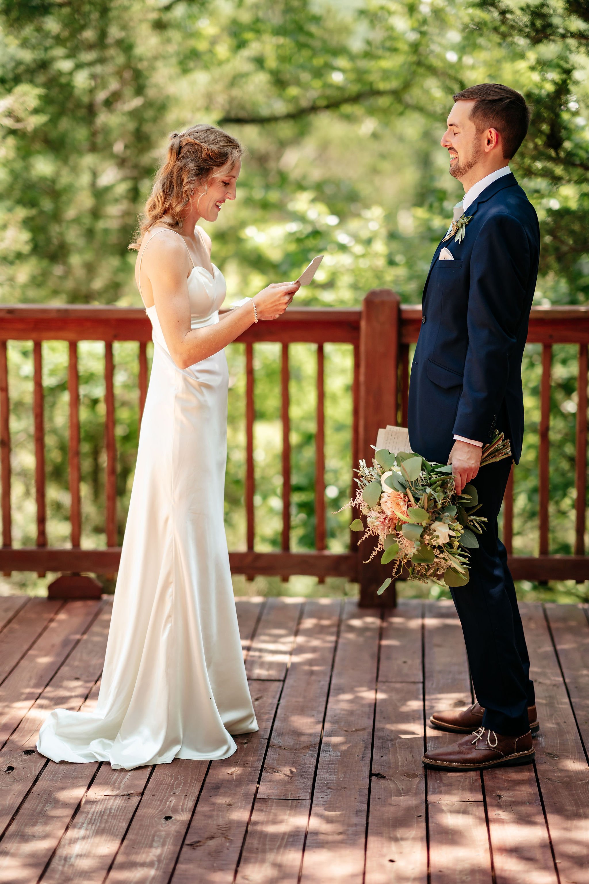 A bride and groom exchanging vows outdoors on a wooden deck surrounded by trees.