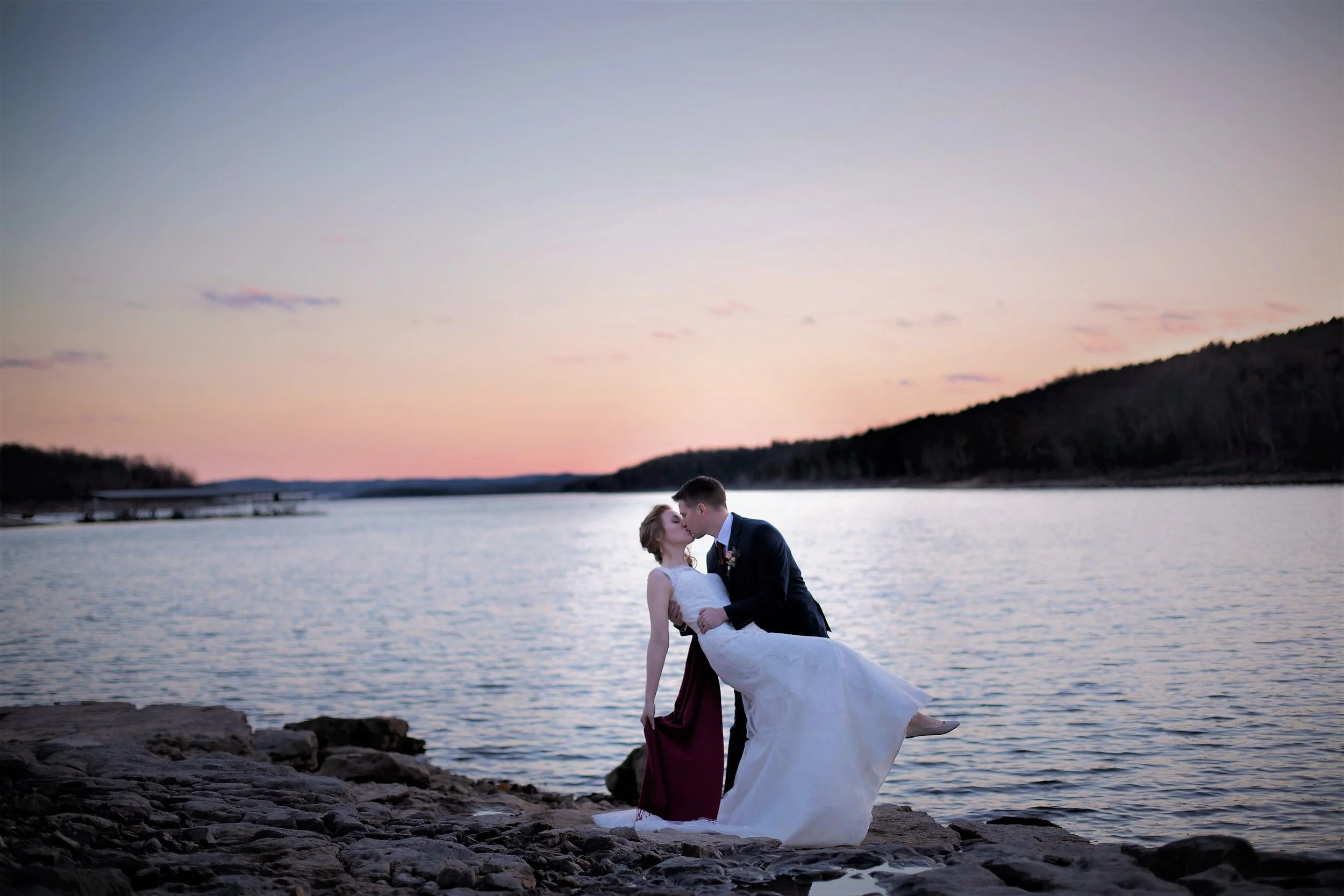 A newlywed couple kissing by a body of water at sunset, with mountains in the background.