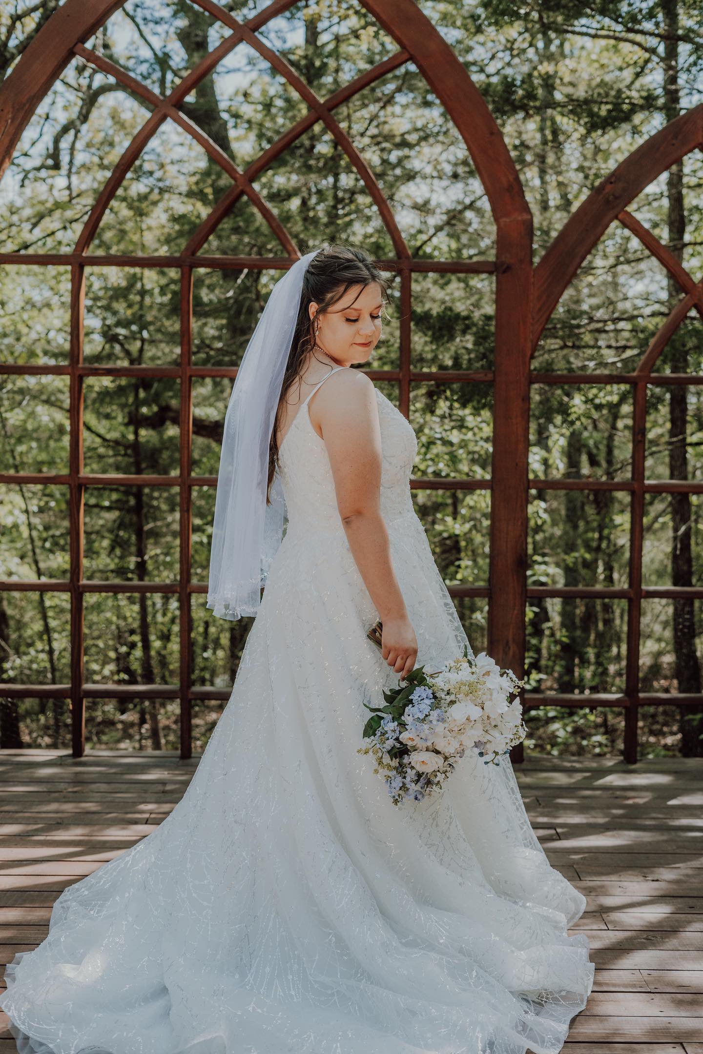 A bride in a white wedding gown holding a bouquet of white and purple flowers, standing outdoors in front of a wooden structure with trees and sunlight in the background.