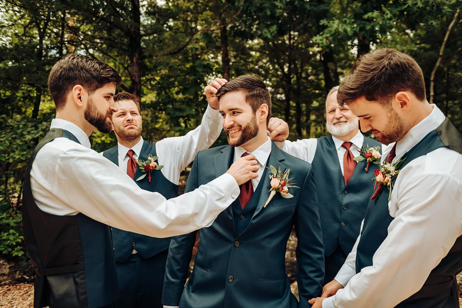 A groom in a navy suit and five groomsmen in vests and white shirts outdoors, with one man adjusting the groom's tie and others smiling or looking down, in a wooded area with trees.