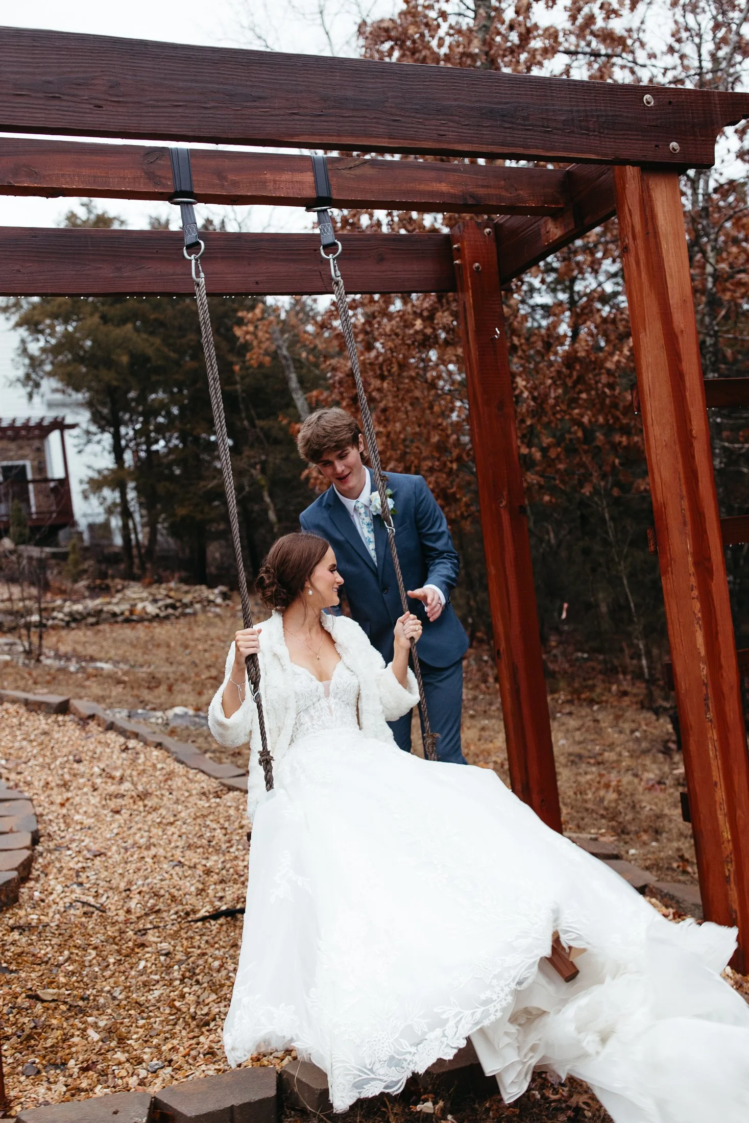 A bride in a white wedding dress and veil sitting on a swing at an outdoor wedding venue while a groom in a blue suit stands behind her, holding the swing's ropes and smiling.