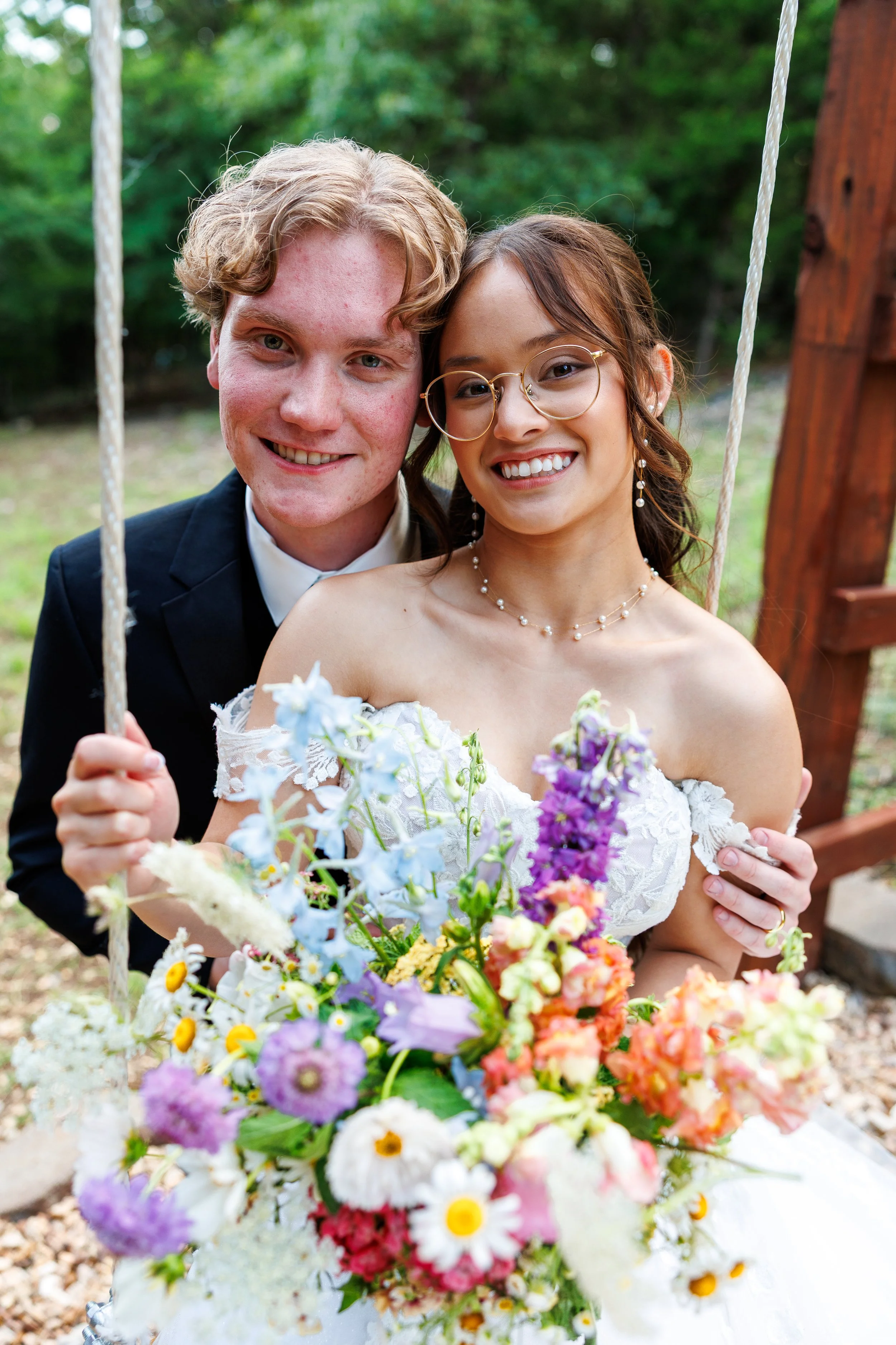 A smiling couple at a wedding, the man in a black tuxedo and the woman in a white wedding dress, holding a bouquet of colorful flowers outdoors with green foliage in the background.