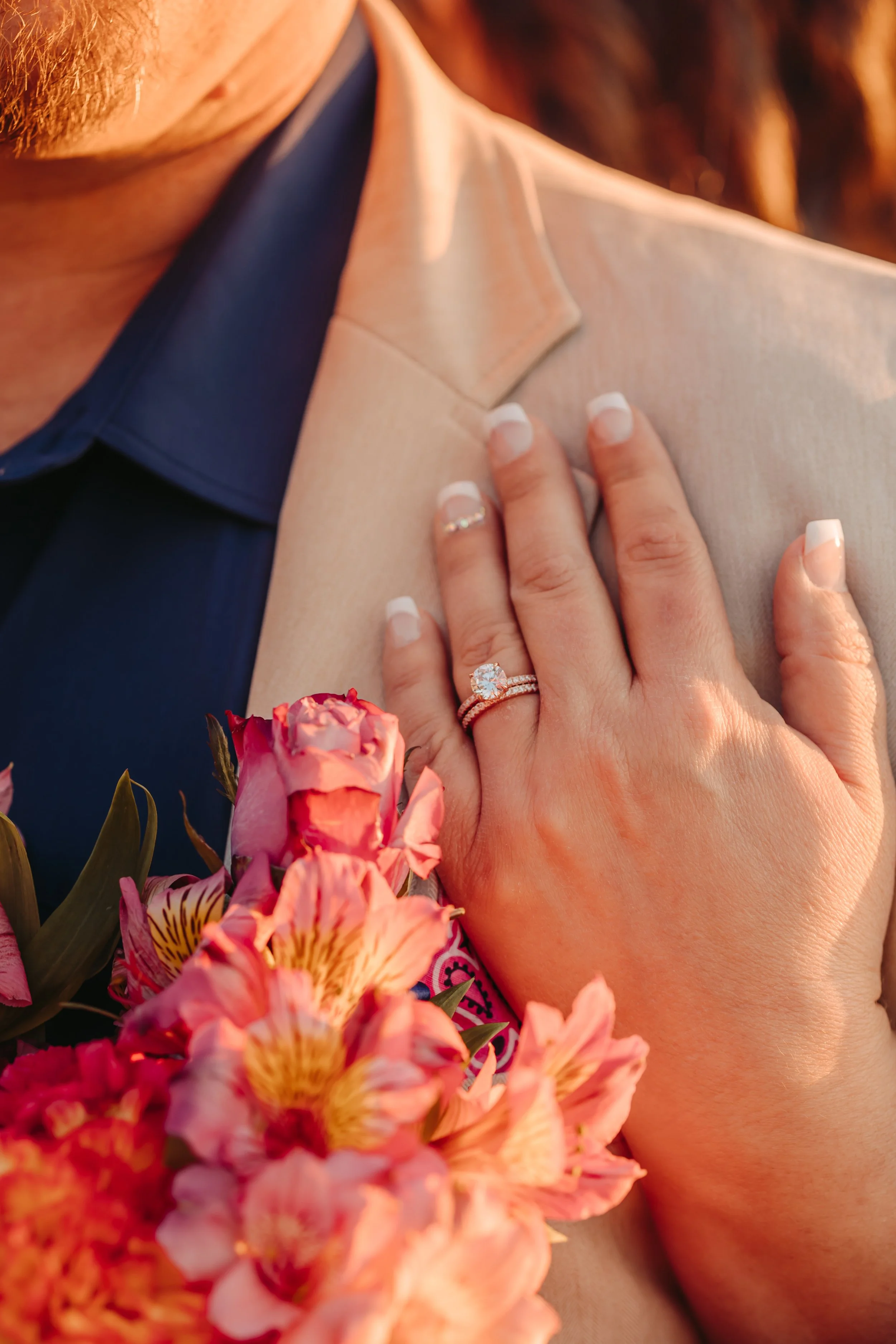 Close-up of a person wearing a beige blazer and blue shirt, touching their chest, with pink flowers in the foreground.