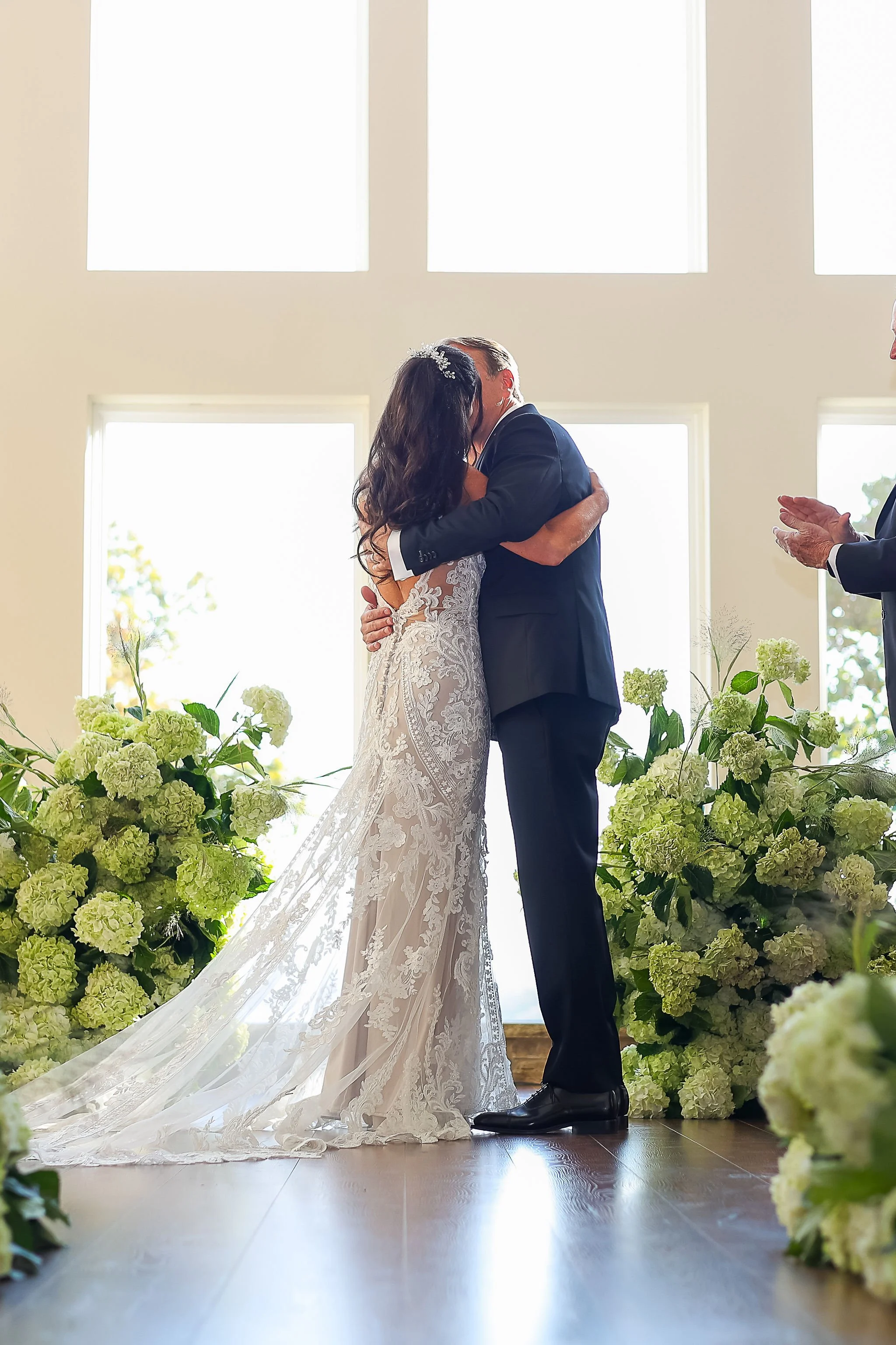 A bride and groom share a kiss at their wedding ceremony in a sunlit room, surrounded by white flower arrangements.