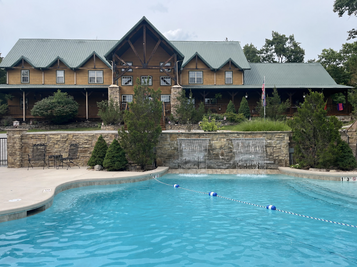 A large outdoor swimming pool with water features, surrounded by a deck and lounge chairs, in front of a rustic multi-story building with a front porch, multiple windows, and a green metal roof, decorated with plants and an American flag.