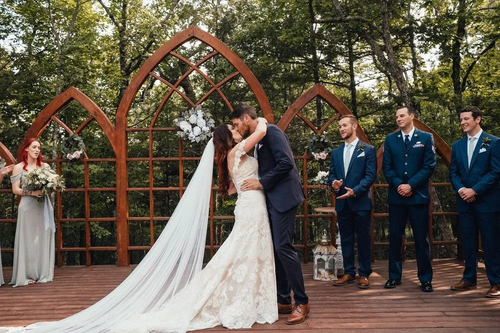 A wedding ceremony outdoors with a bride and groom kissing on a wooden deck, surrounded by wedding party members in formal attire, with forest in the background.