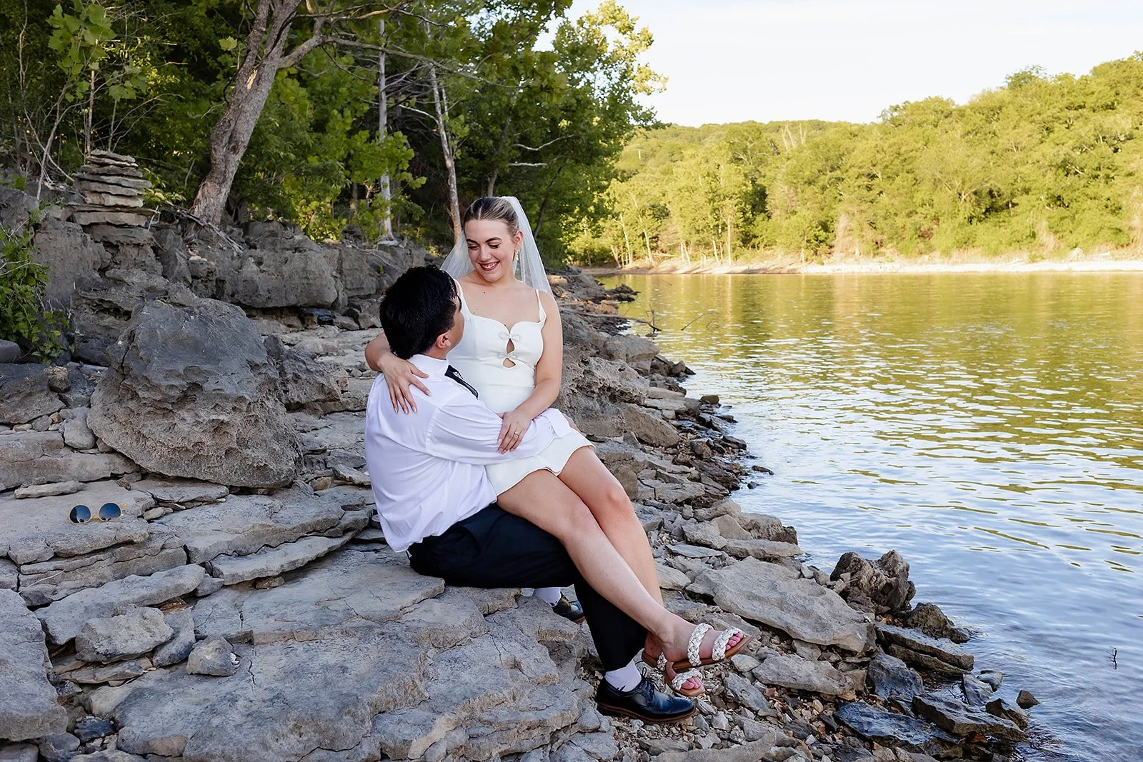 A couple sitting on rocks by a river, with green trees and a clear sky in the background. The woman is wearing a white dress and a veil, smiling, while the man is wearing a white shirt and black pants, holding her on his lap.