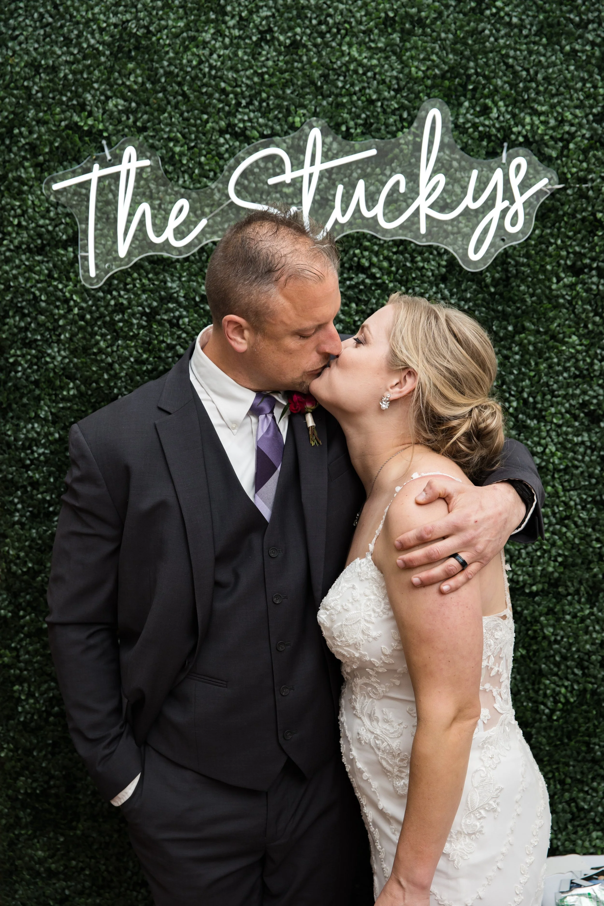 A couple sharing a kiss during their wedding, standing in front of a green hedge with a neon sign that reads 'The Stuckys' in cursive.