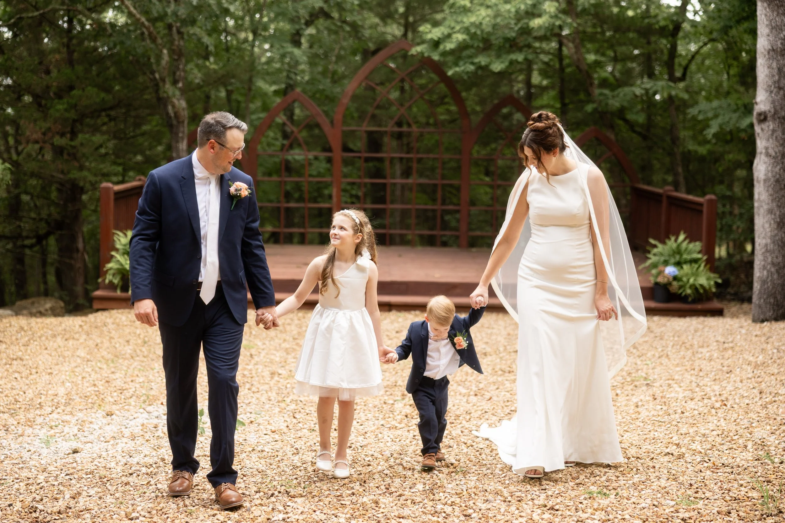 A bride and groom holding hands with two small children during a wedding ceremony outdoors in a wooded area, with a wooden arch in the background.