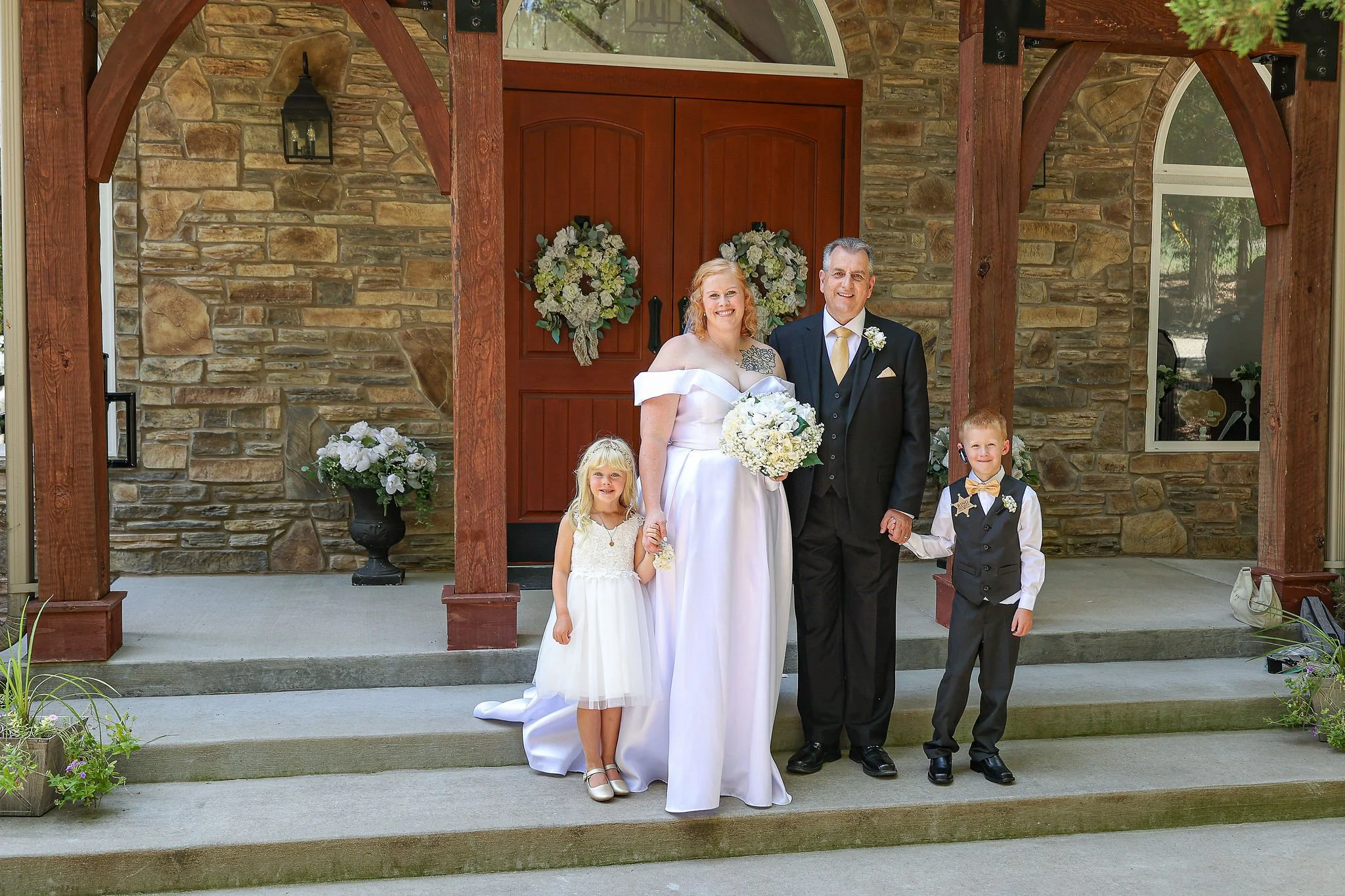 A bride and groom with two young children standing on the steps of a house with a stone facade and wooden porch structure, decorated with floral arrangements and wreaths for a wedding.