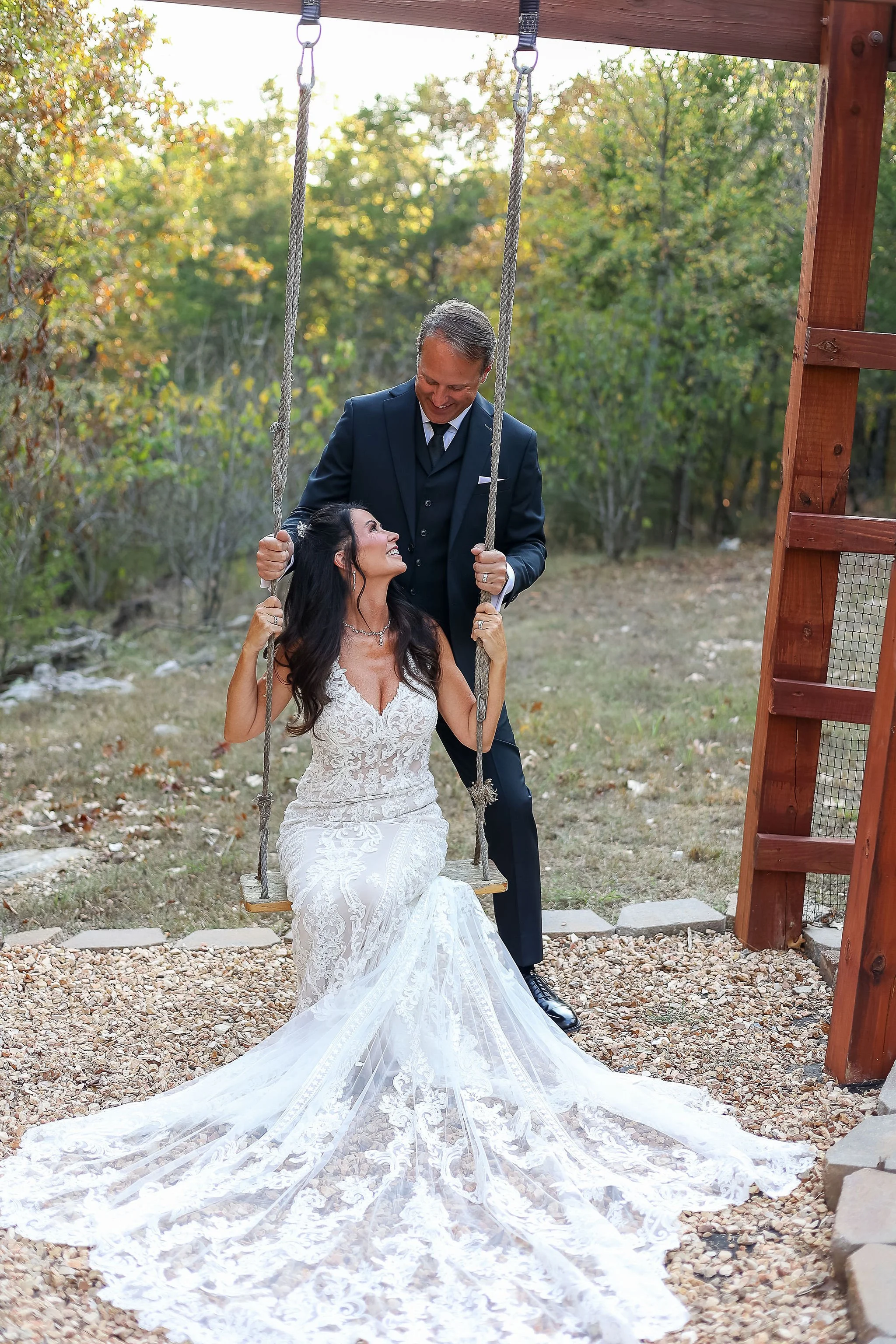 A bride in a white lace wedding dress sitting on a wooden swing, looking up at a groom in a dark suit standing beside her, outdoors in a natural setting with trees and sunlight in the background.