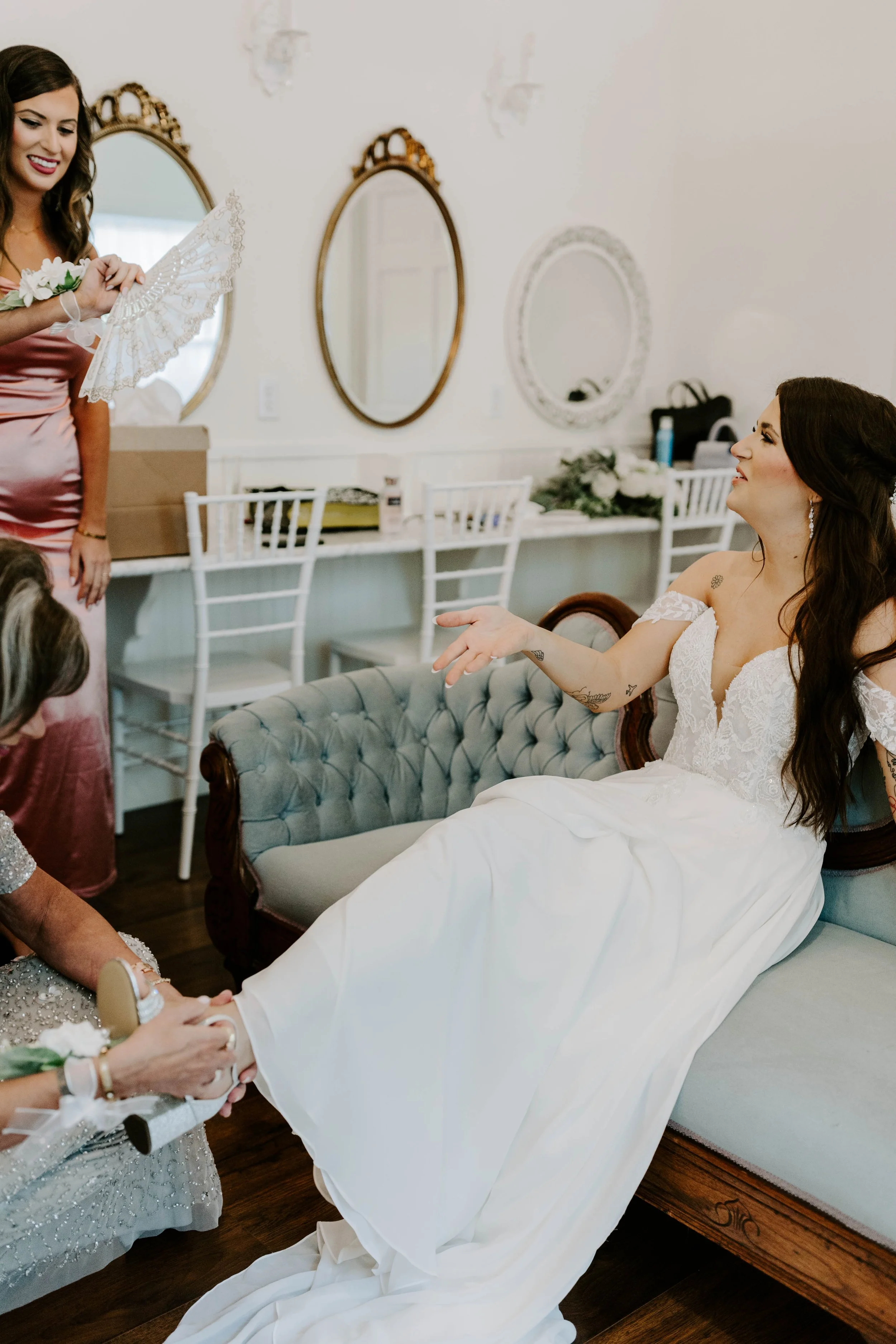 A bride in a white wedding dress is seated on a vintage-style sofa, talking to a woman in a pink satin dress who is holding her foot. Another woman in a sequined dress is assisting her with her shoes. The background has a row of framed mirrors, a dec
