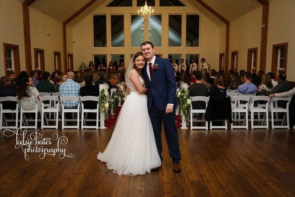 A bride and groom pose together during their wedding reception in a decorated hall, with guests seated in chairs behind them and a band or musicians on stage in the background.
