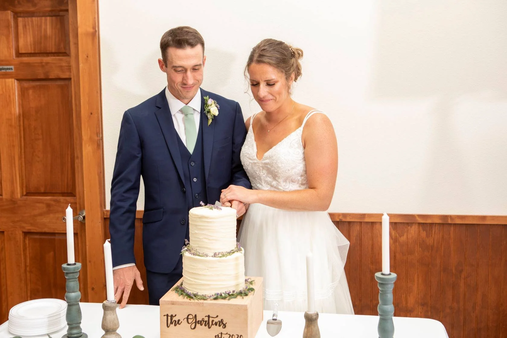 A bride and groom cutting a wedding cake at their reception. The bride is wearing a white dress, and the groom is in a dark blue suit with a light green tie. There are lit candles on the table and a wooden wall in the background.