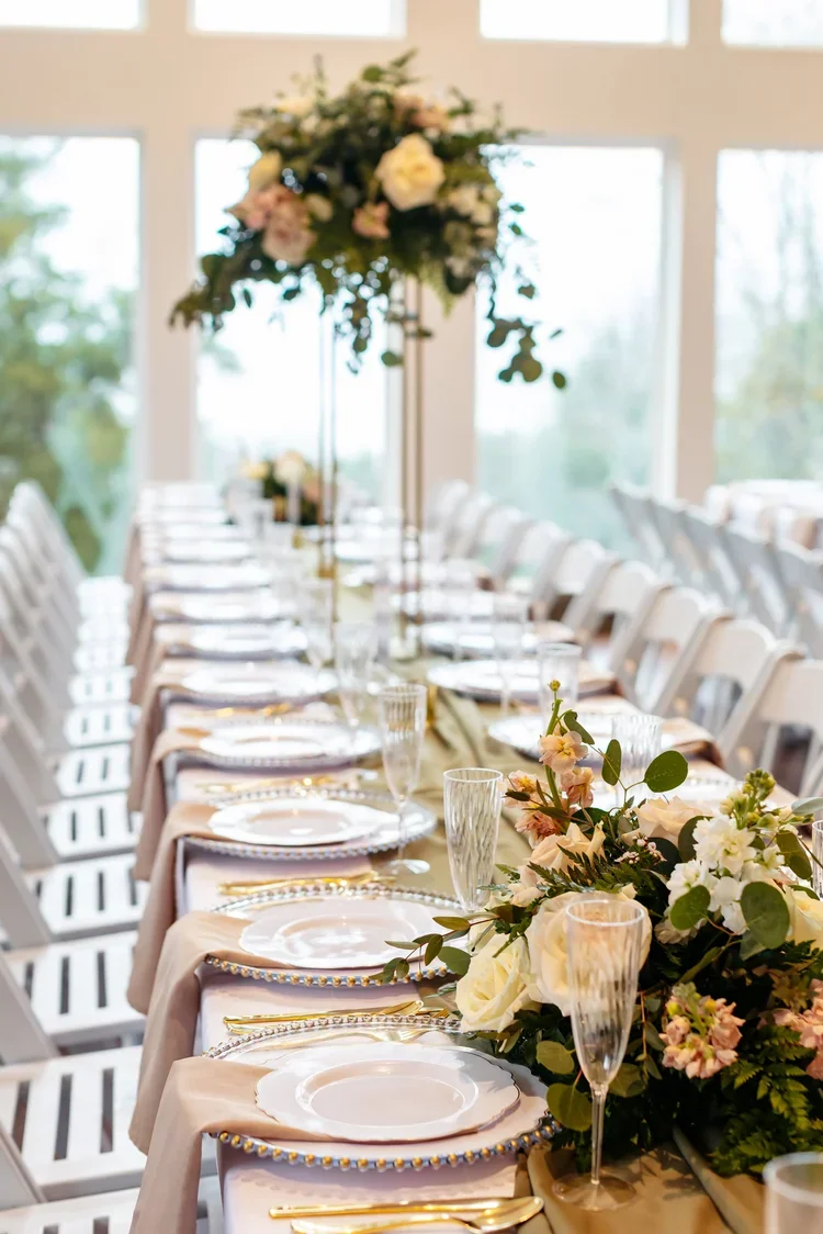 Elegant banquet table setting with floral centerpieces, gold-rimmed plates, gold utensils, and glassware in a bright room with large windows.